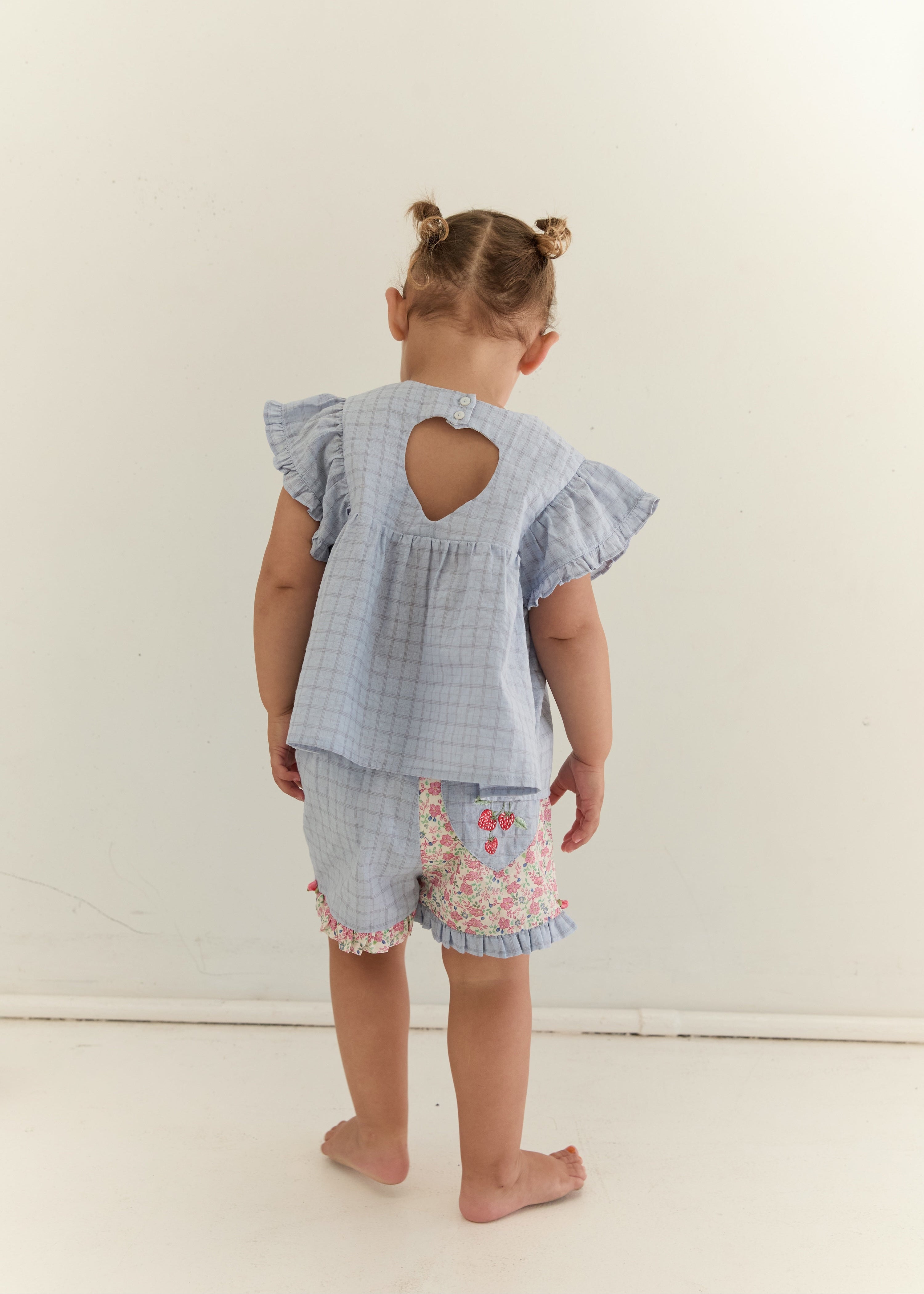 A toddler standing with their back to the camera, wearing a light blue and white gingham two-piece outfit with a ruffled back cutout on the top and floral trim on the shorts, set against a plain off-white wall.