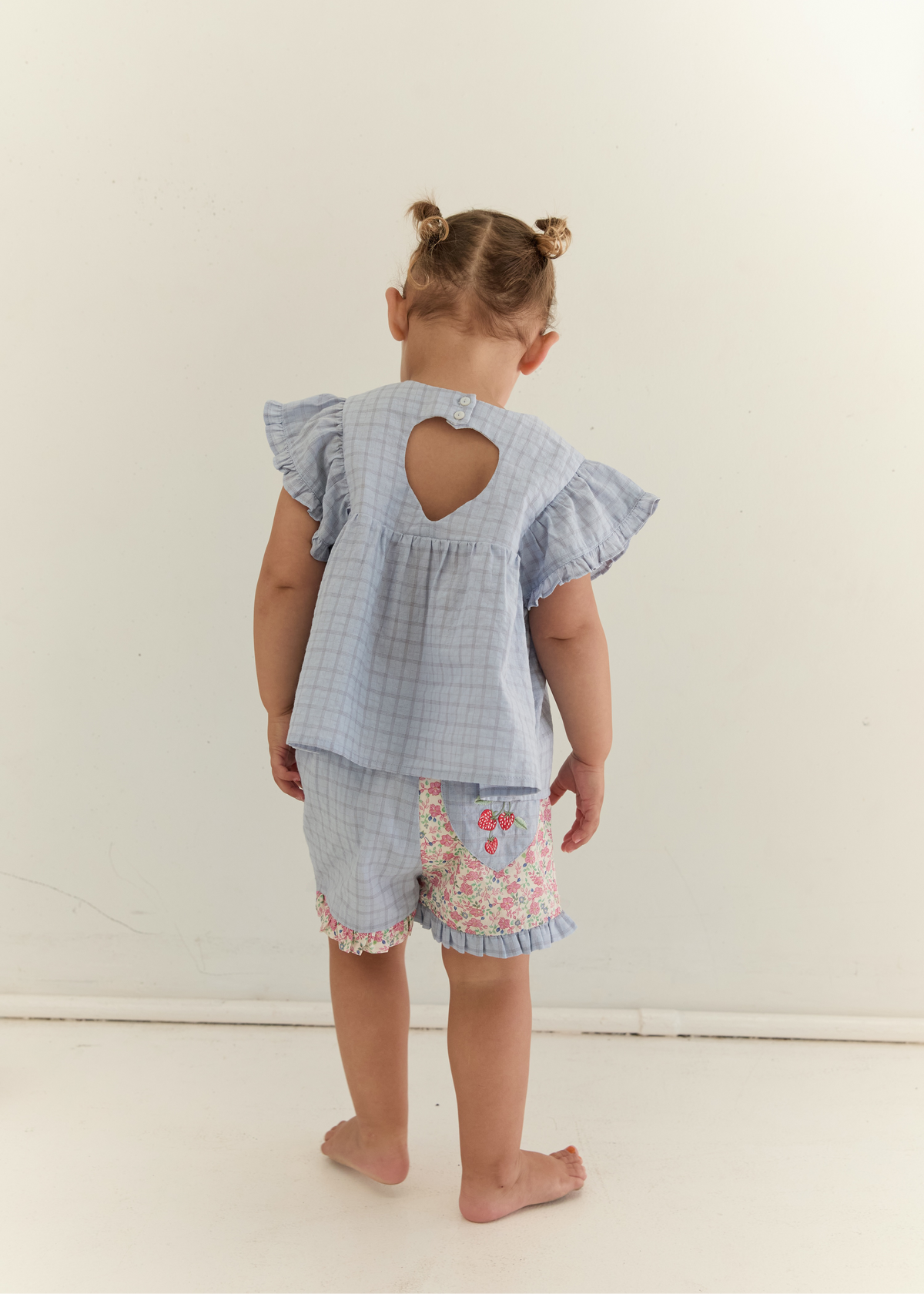 A toddler standing with their back to the camera, wearing a light blue and white gingham two-piece outfit with a ruffled back cutout on the top and floral trim on the shorts, set against a plain off-white wall.
