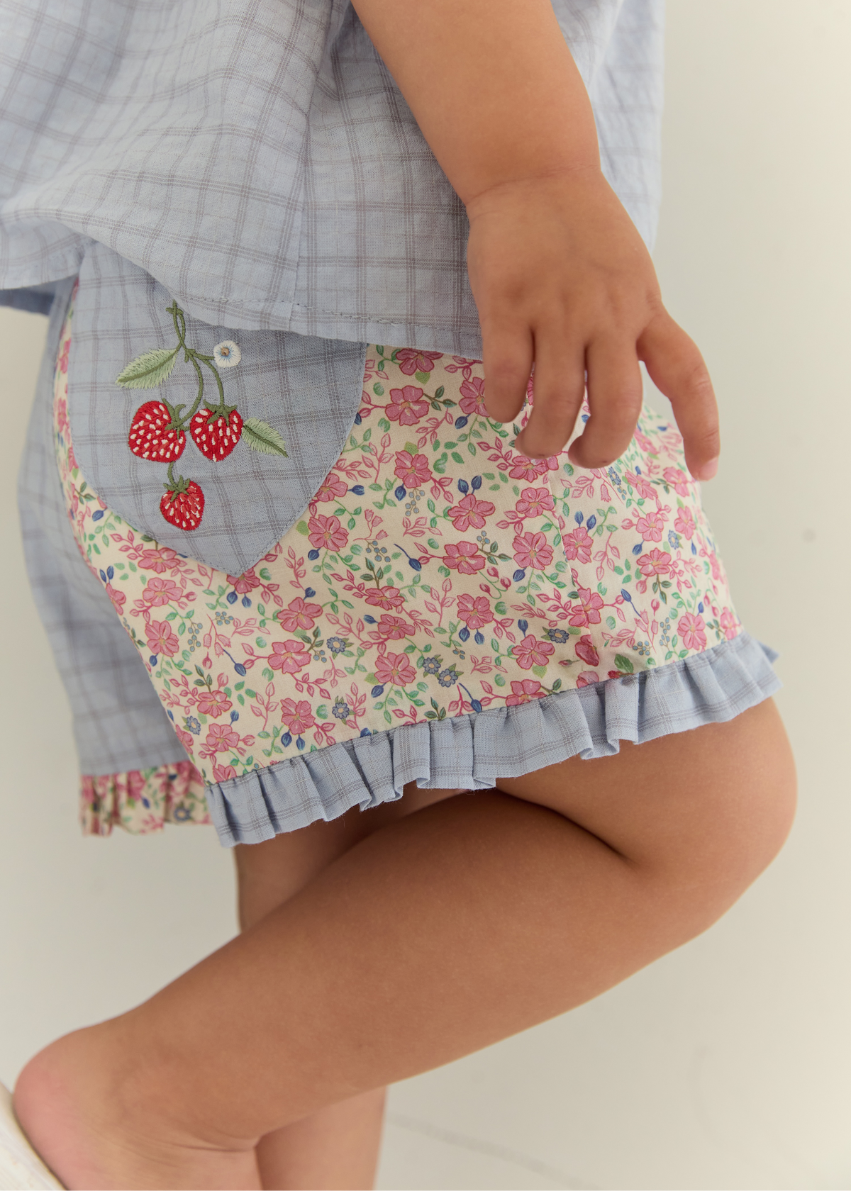 Close-up of a child wearing a light blue plaid skirt with a floral patch pocket and embroidered red strawberries on the side.