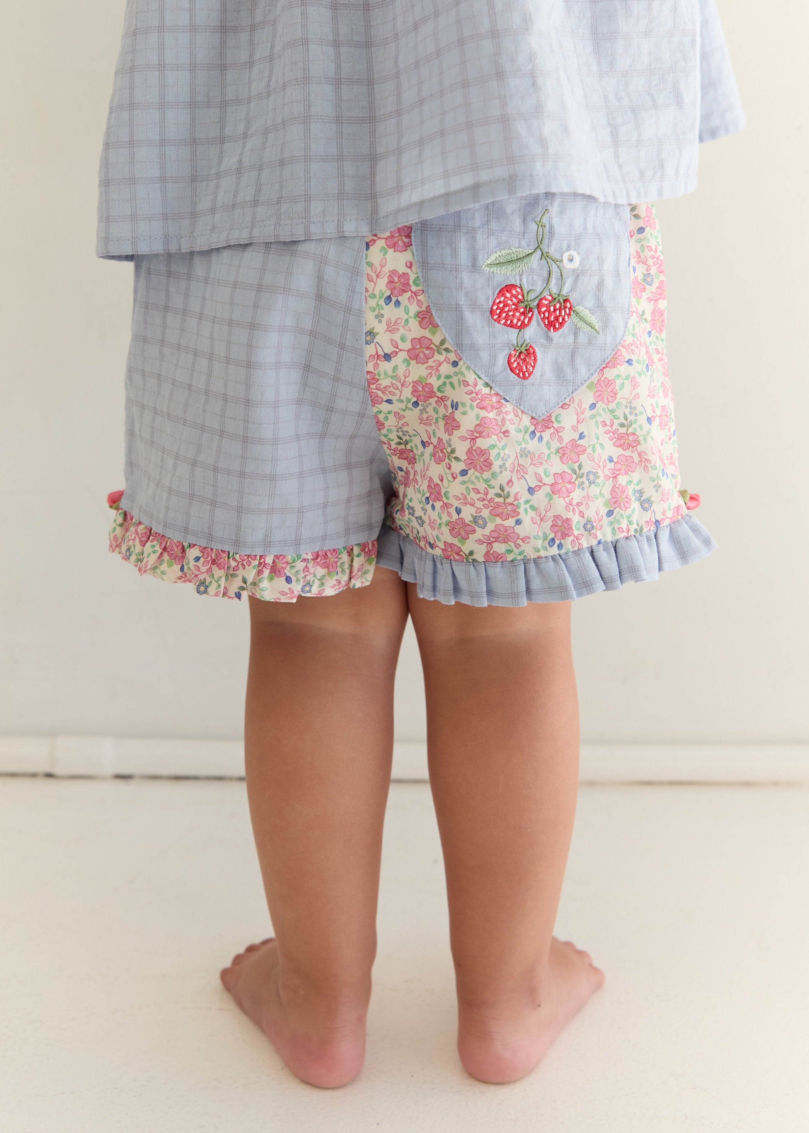 A child shown from the back wearing light blue plaid shorts with a floral patch pocket featuring embroidered cherries, paired with a matching light blue plaid top. The child is barefoot on a light beige floor against a neutral wall.