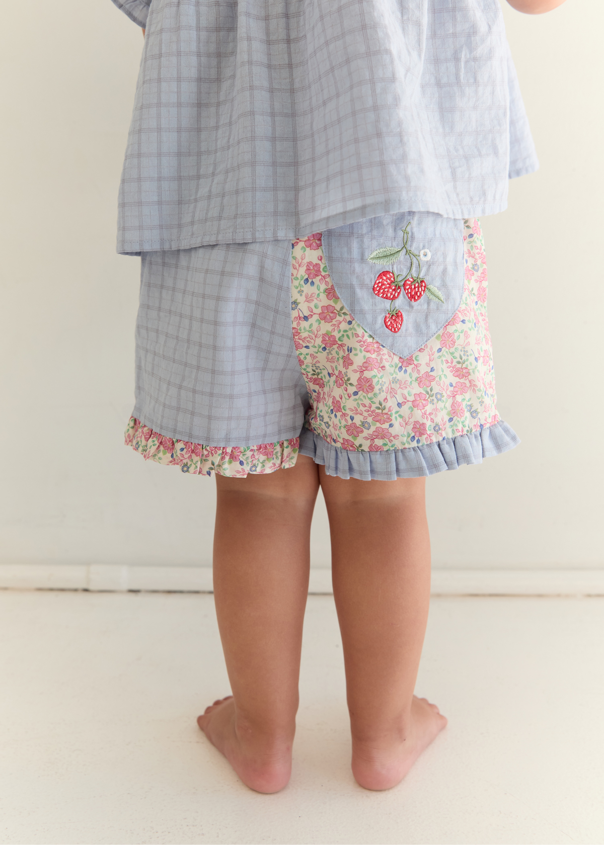 A child shown from the back wearing light blue plaid shorts with a floral patch pocket featuring embroidered cherries, paired with a matching light blue plaid top. The child is barefoot on a light beige floor against a neutral wall.