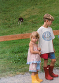 Three children standing outdoors in a mountain meadow with a waterfall and rocky cliff in the background. The children are wearing casual summer clothes and colorful rain boots.