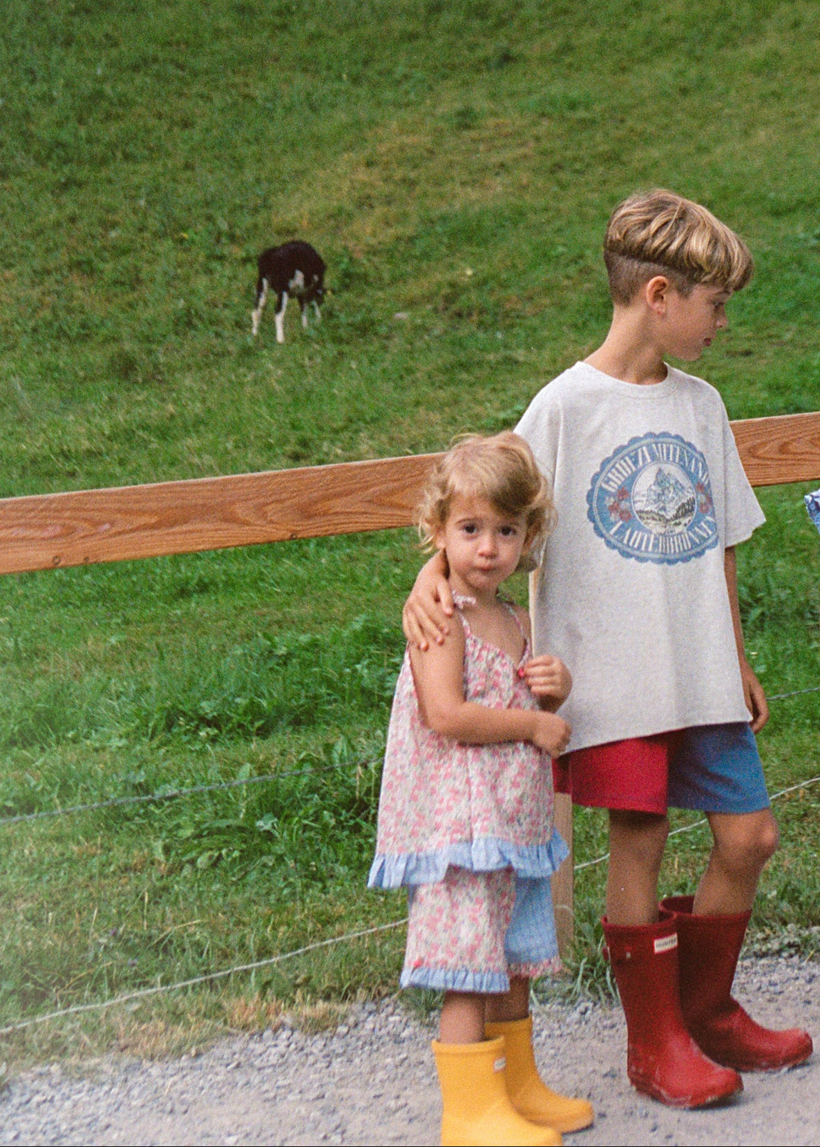 Three children standing outdoors in a mountain meadow with a waterfall and rocky cliff in the background. The children are wearing casual summer clothes and colorful rain boots.