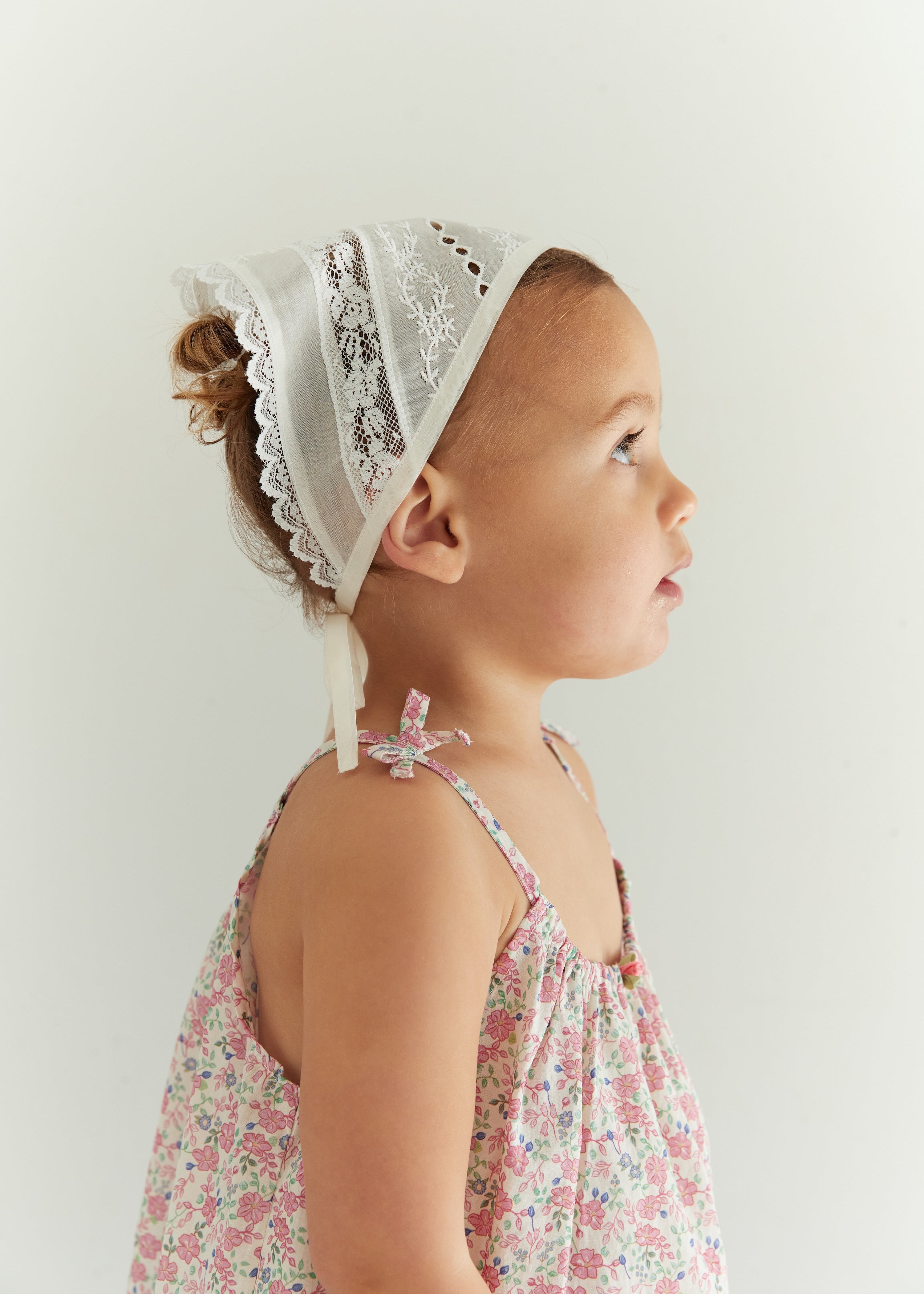 A toddler girl in profile wearing a white lace-trim bonnet and a pink floral spaghetti-strap dress, set against a plain light gray background.