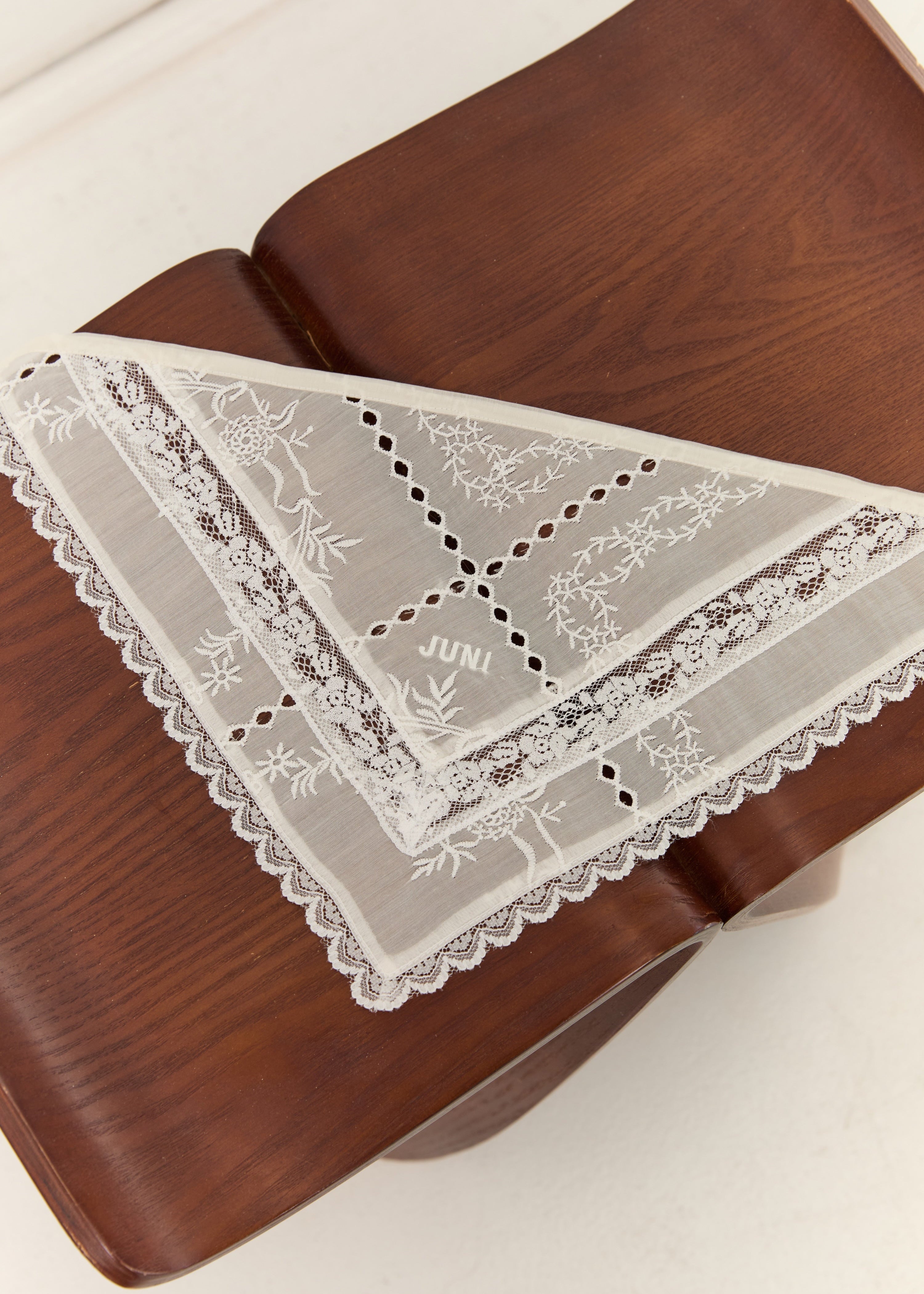 A white lace square handkerchief with floral embroidery and scalloped edges, folded and draped on a dark brown wooden surface.