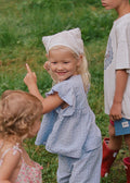 Three children standing on a grassy field wearing matching light blue gingham rain suit and red rain boots, with one child pointing upward.