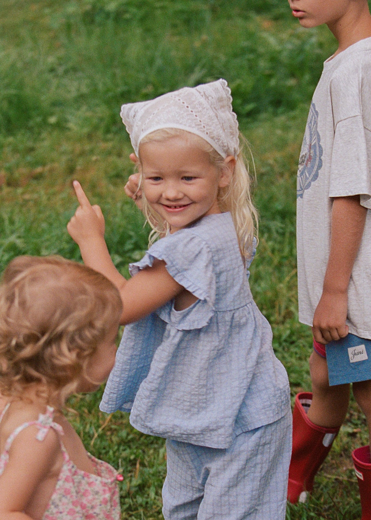 Three children standing on a grassy field wearing matching light blue gingham rain suit and red rain boots, with one child pointing upward.