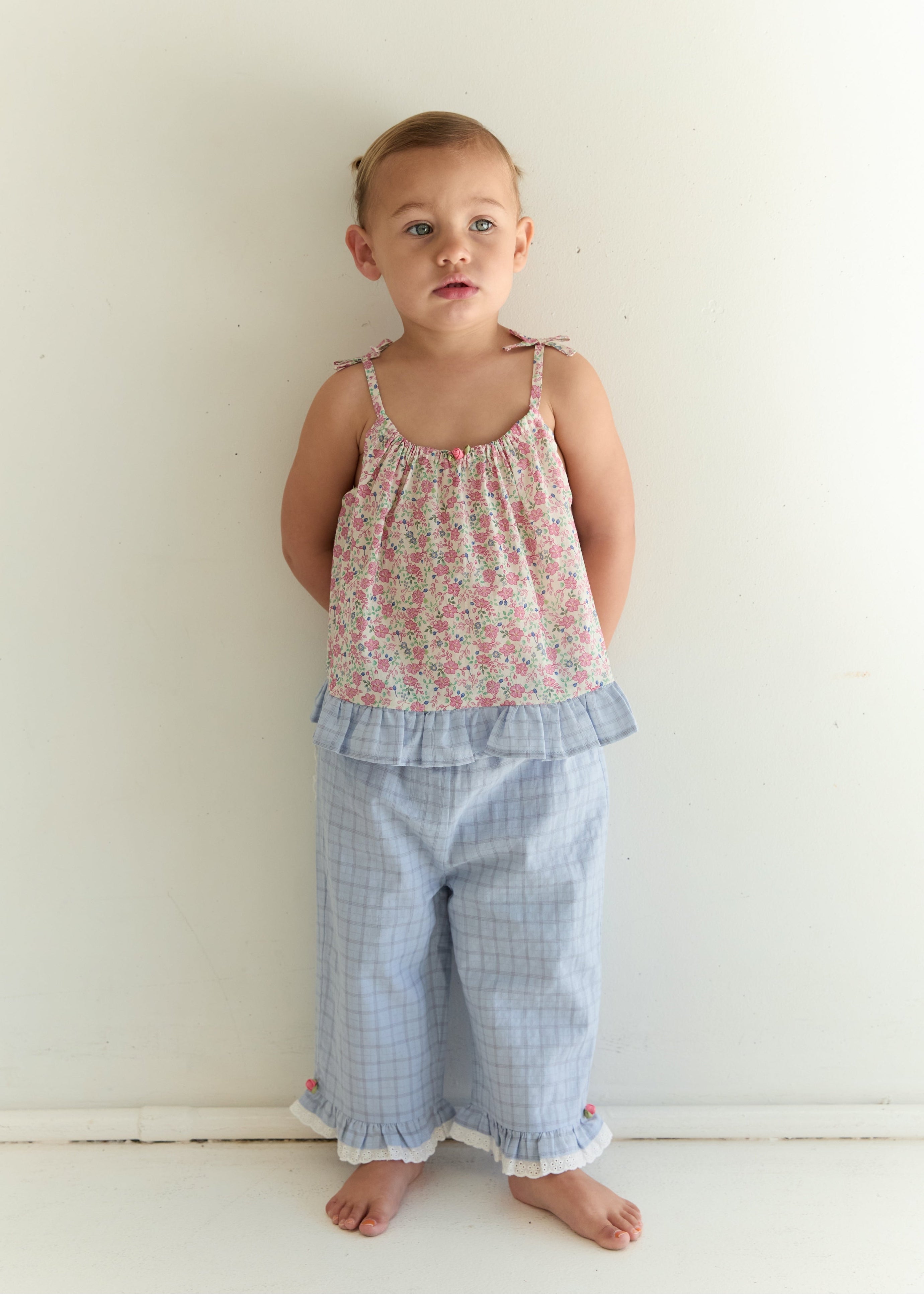 A toddler stands against a plain white wall wearing a two-piece pajama set with a pink floral camisole top and light blue plaid pants with ruffled trim.