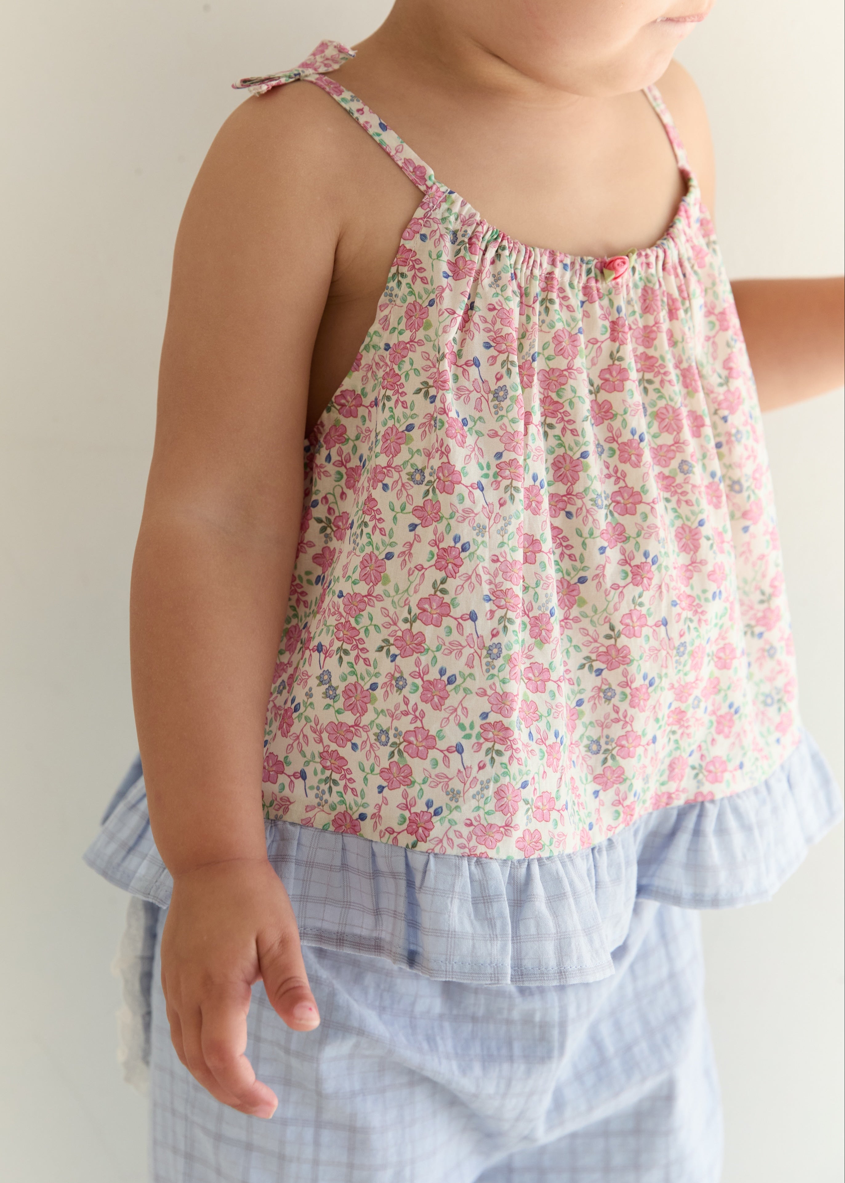 A toddler wearing a sleeveless floral camisole top with thin shoulder ties and a ruffled hem, paired with light blue plaid shorts, photographed against a plain light background.