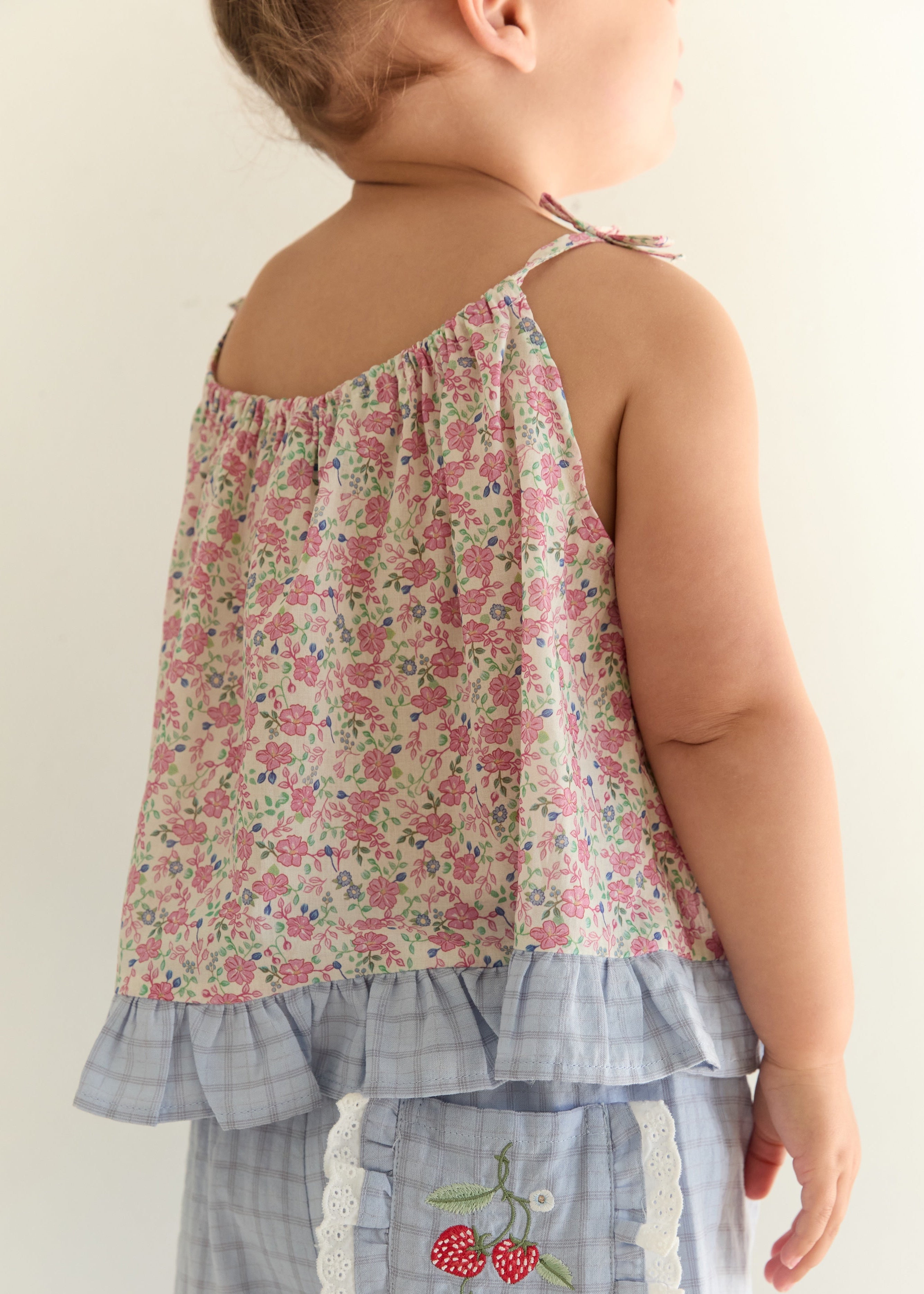 A toddler shown from the back wearing a sleeveless floral top with ruffled trim and light blue plaid pants featuring a cherry applique pocket.