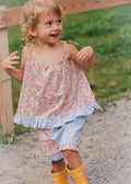 A young girl smiling outdoors, wearing a pink and white floral sleeveless top with a ruffled hem, matching floral shorts, and bright yellow rain boots.