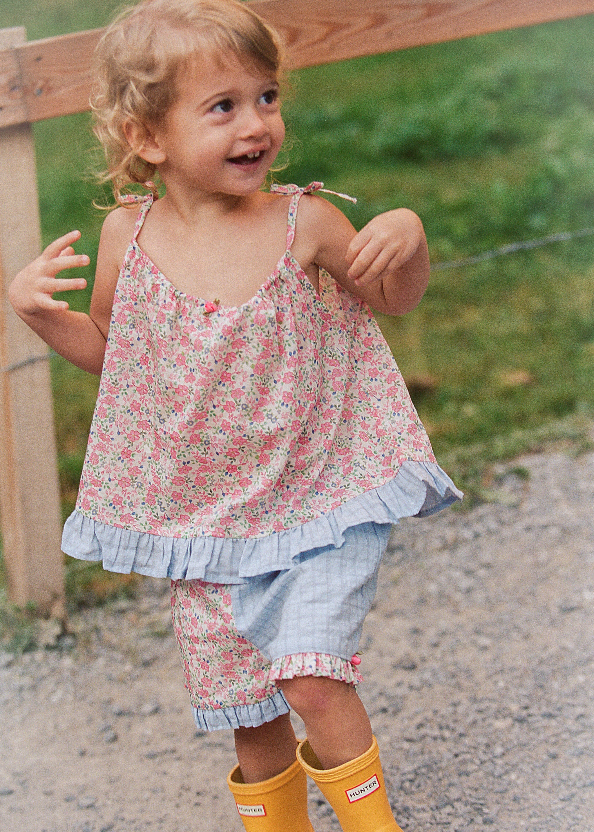 A young girl smiling outdoors, wearing a pink and white floral sleeveless top with a ruffled hem, matching floral shorts, and bright yellow rain boots.