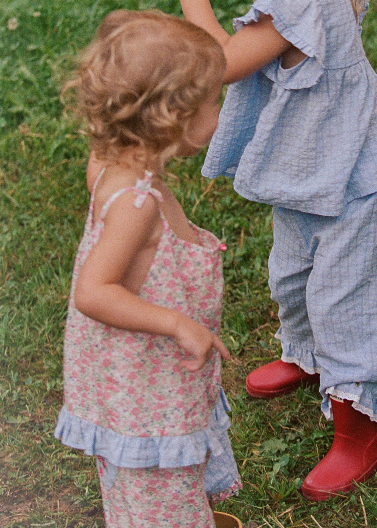 Three children standing on a grassy field wearing matching light blue gingham rain suit and red rain boots, with one child pointing upward.