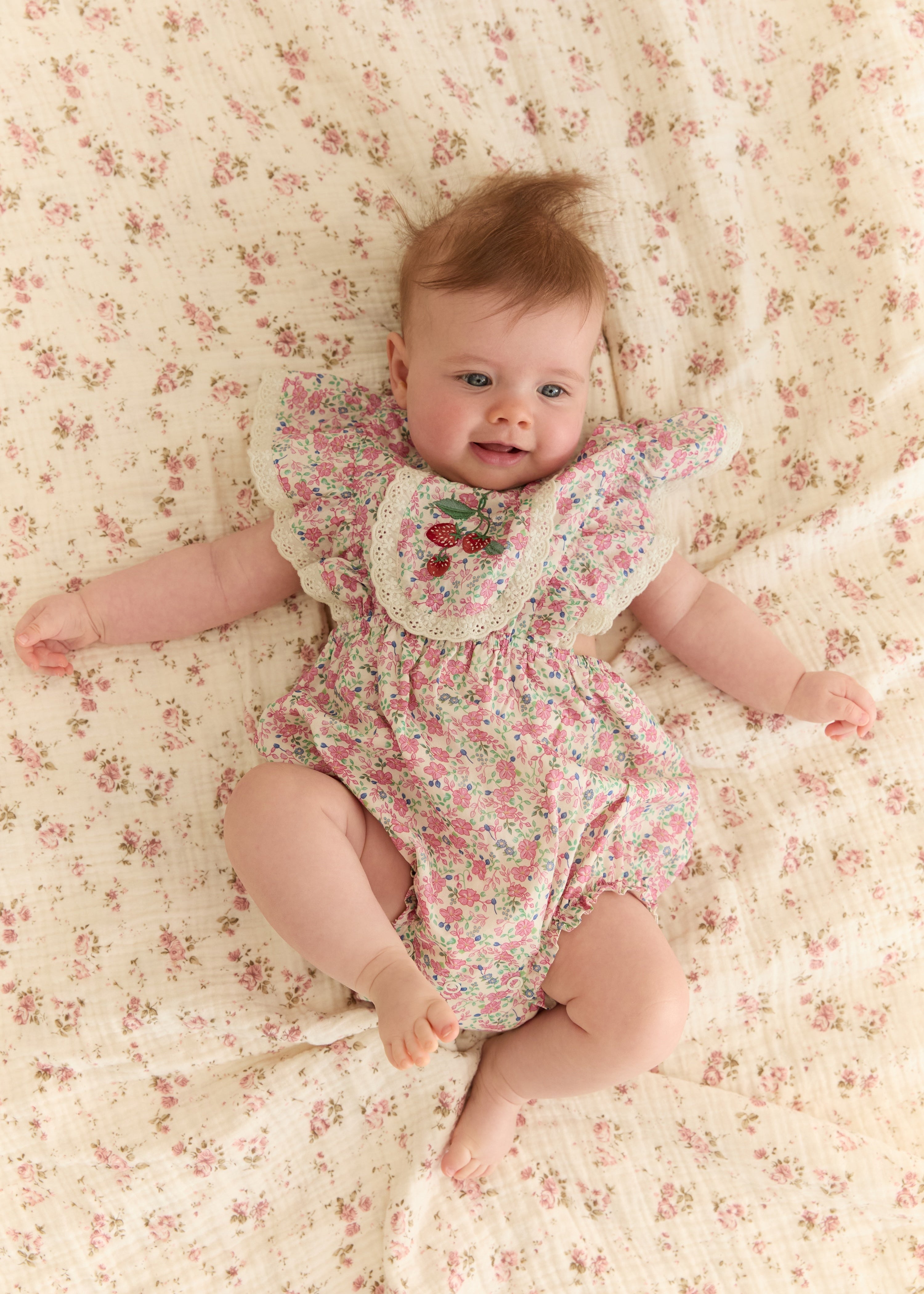 A baby lying on a cream-colored floral blanket, wearing a matching short-sleeve floral romper with a ruffled collar and embroidered cherry details.