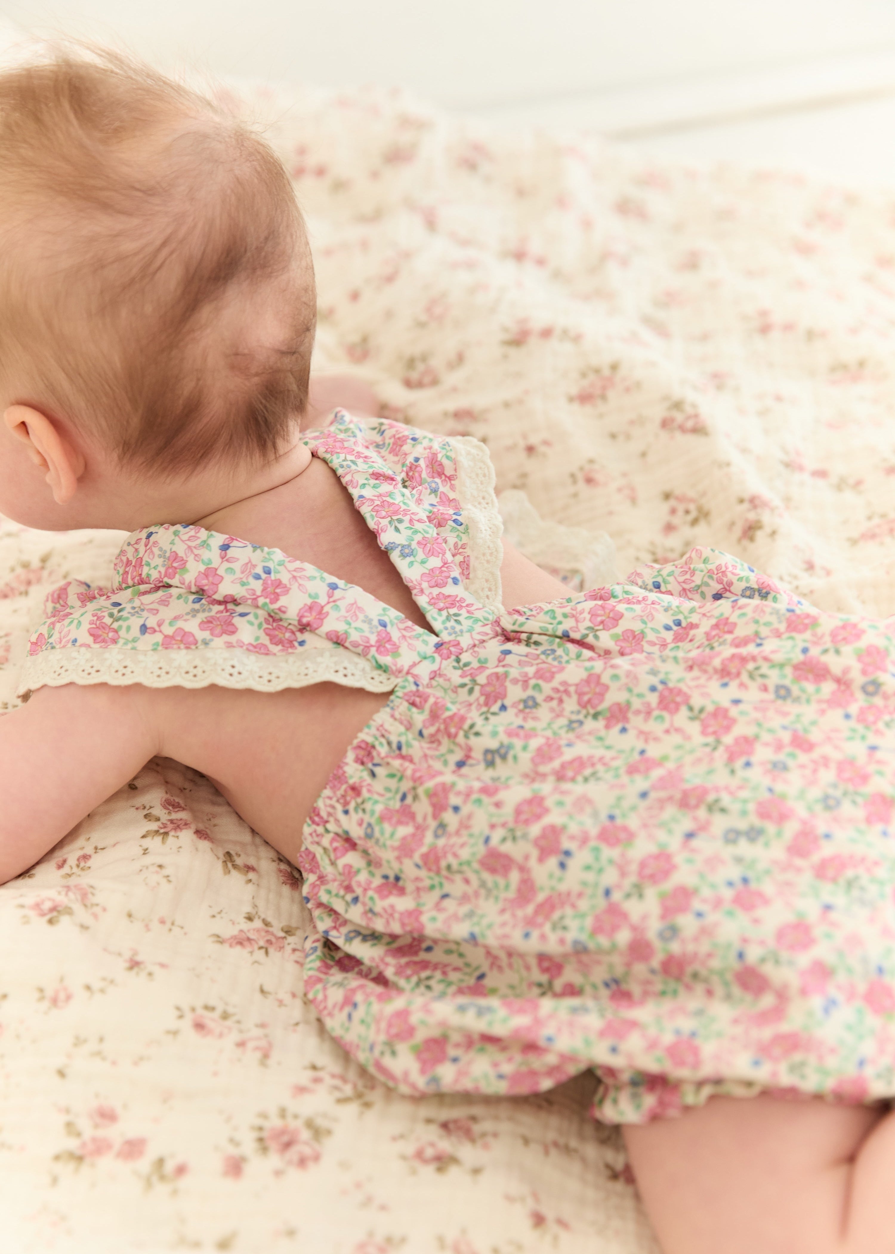 A baby lying on their stomach wearing a pink and green floral romper, photographed in a soft, warm-toned setting.