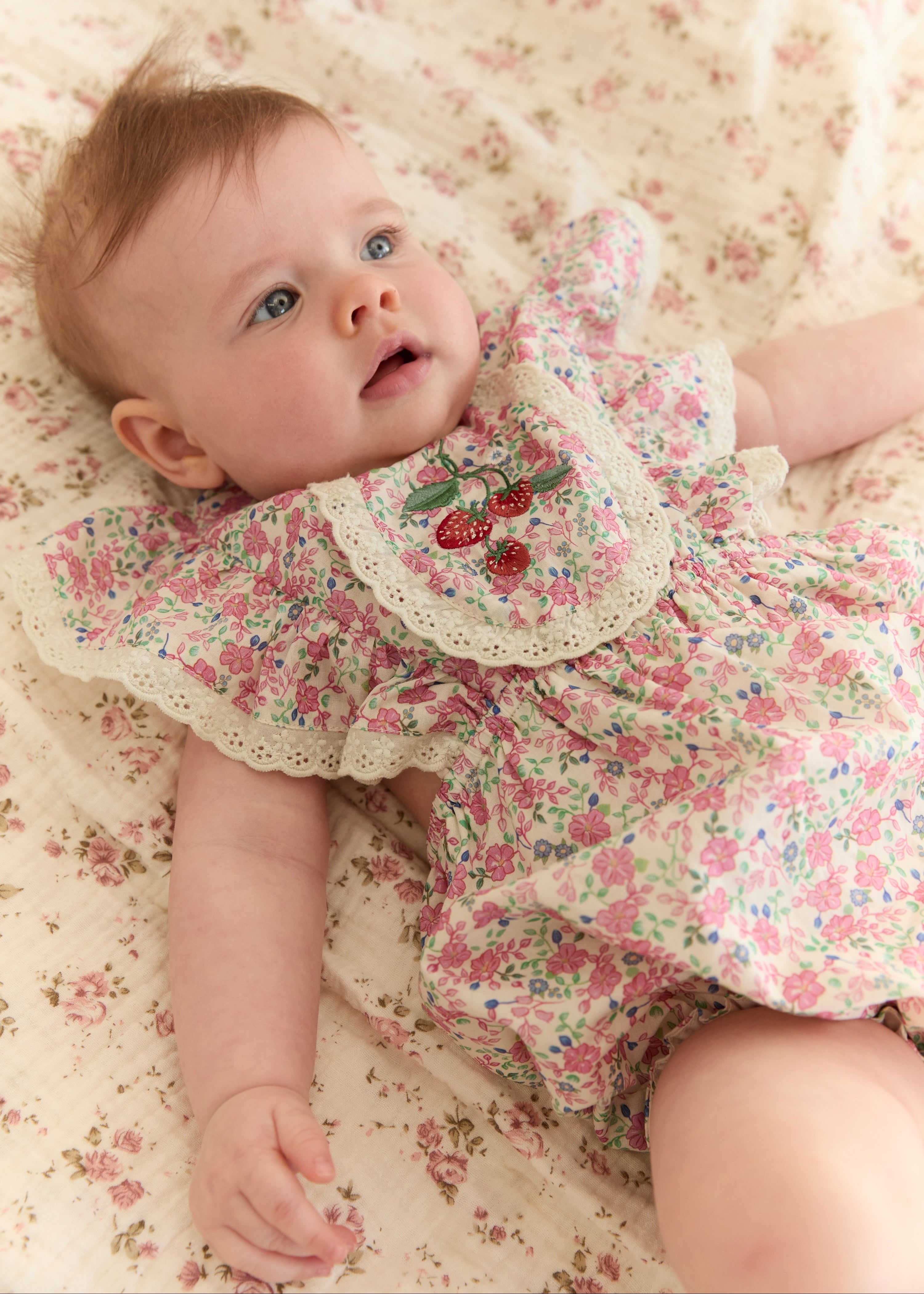 A baby lying on a floral blanket, wearing a matching pink and green floral romper with a white lace collar and embroidered cherries.