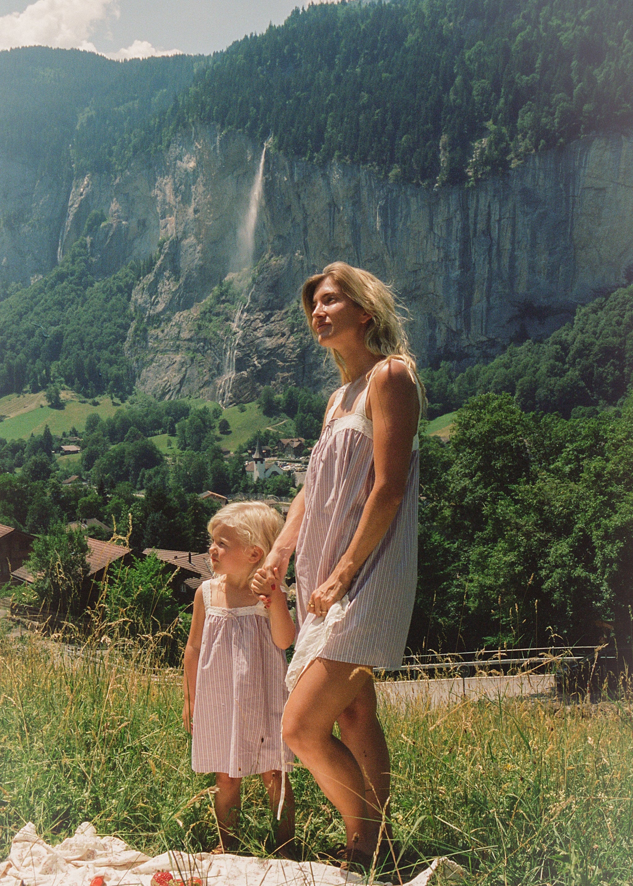 A woman and a young child wearing matching light pink and white striped sleeveless dresses stand on a grassy hillside holding hands, with a tall forested cliff and waterfall in the background.