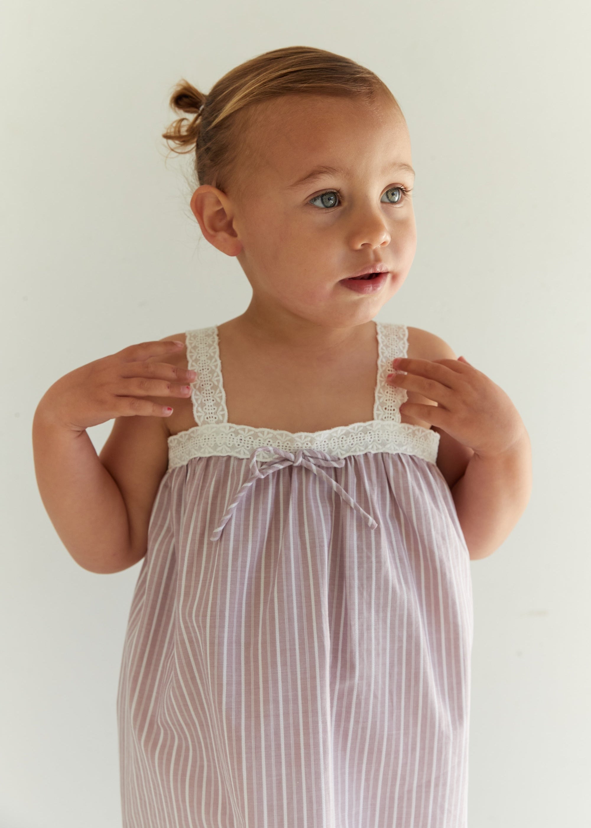 A toddler girl with her hair in a small topknot is wearing a pale pink and white vertically striped nightgown with white lace trim on the straps and neckline. She is posing against a plain, light off-white background.