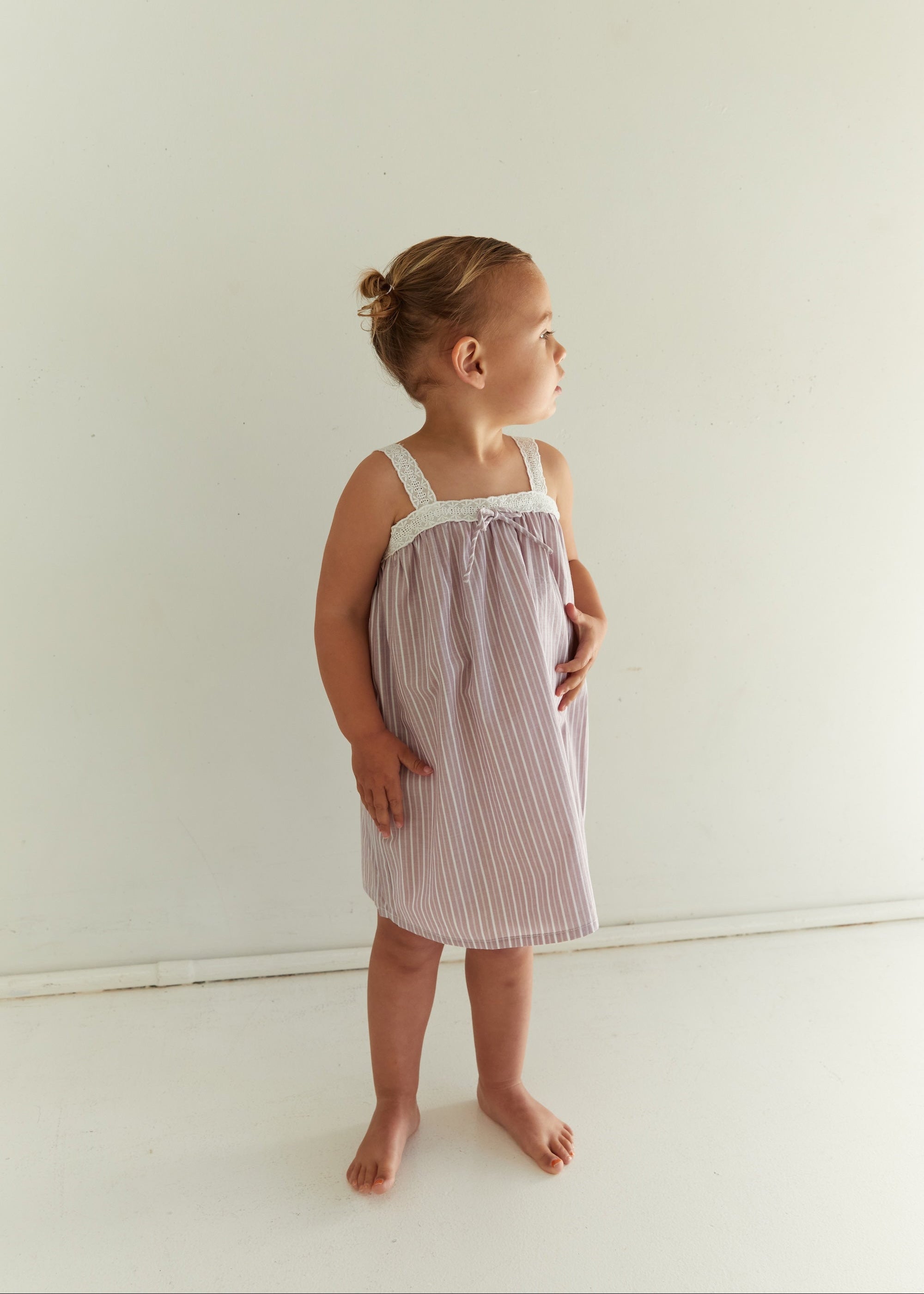 A toddler girl wearing a sleeveless, tie-front nightgown with thin vertical stripes in dusty pink and white, standing against a plain off-white wall.
