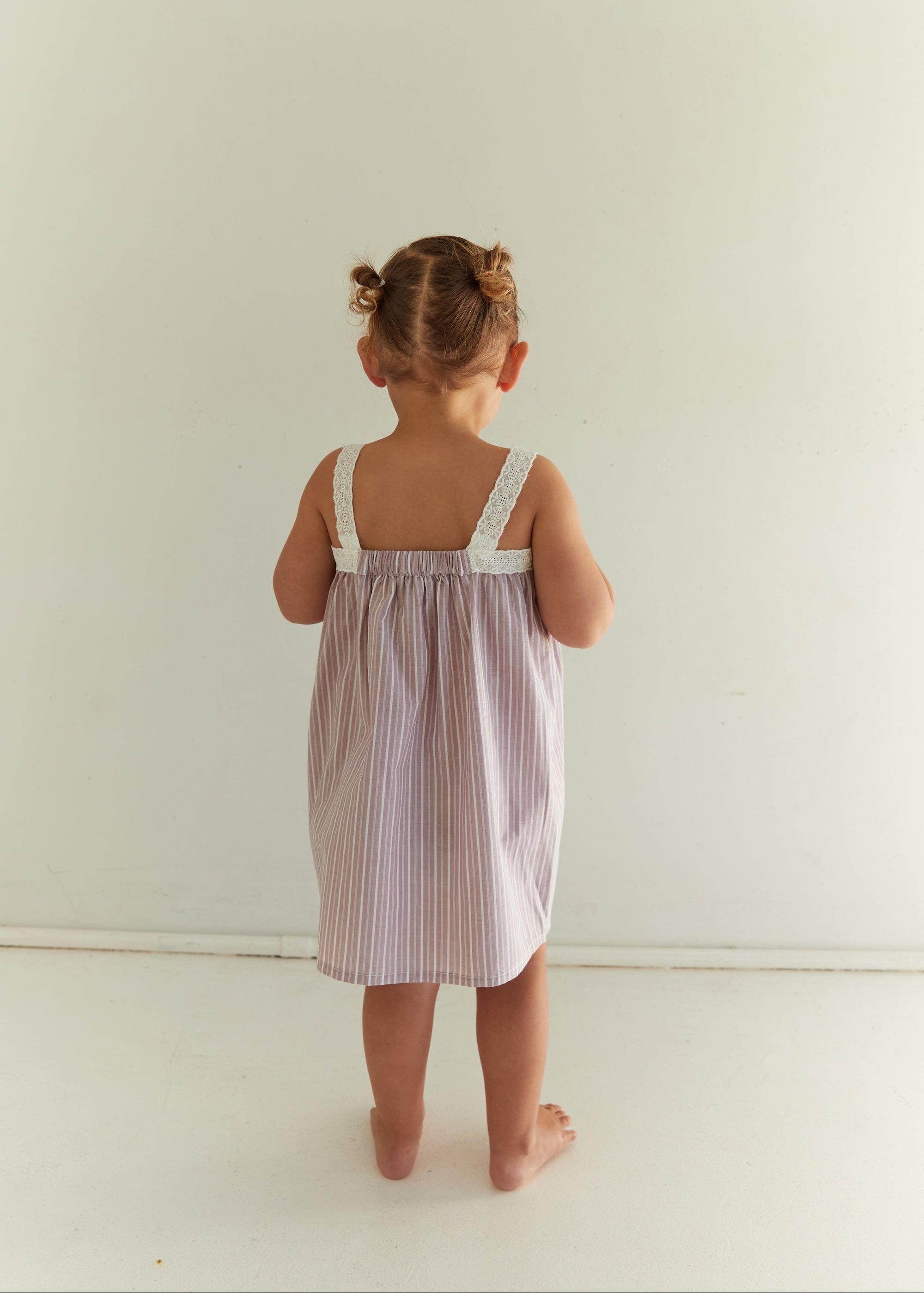 A toddler girl viewed from behind wearing a lavender and white vertically striped nightgown with white lace shoulder straps, standing against a plain off-white wall.