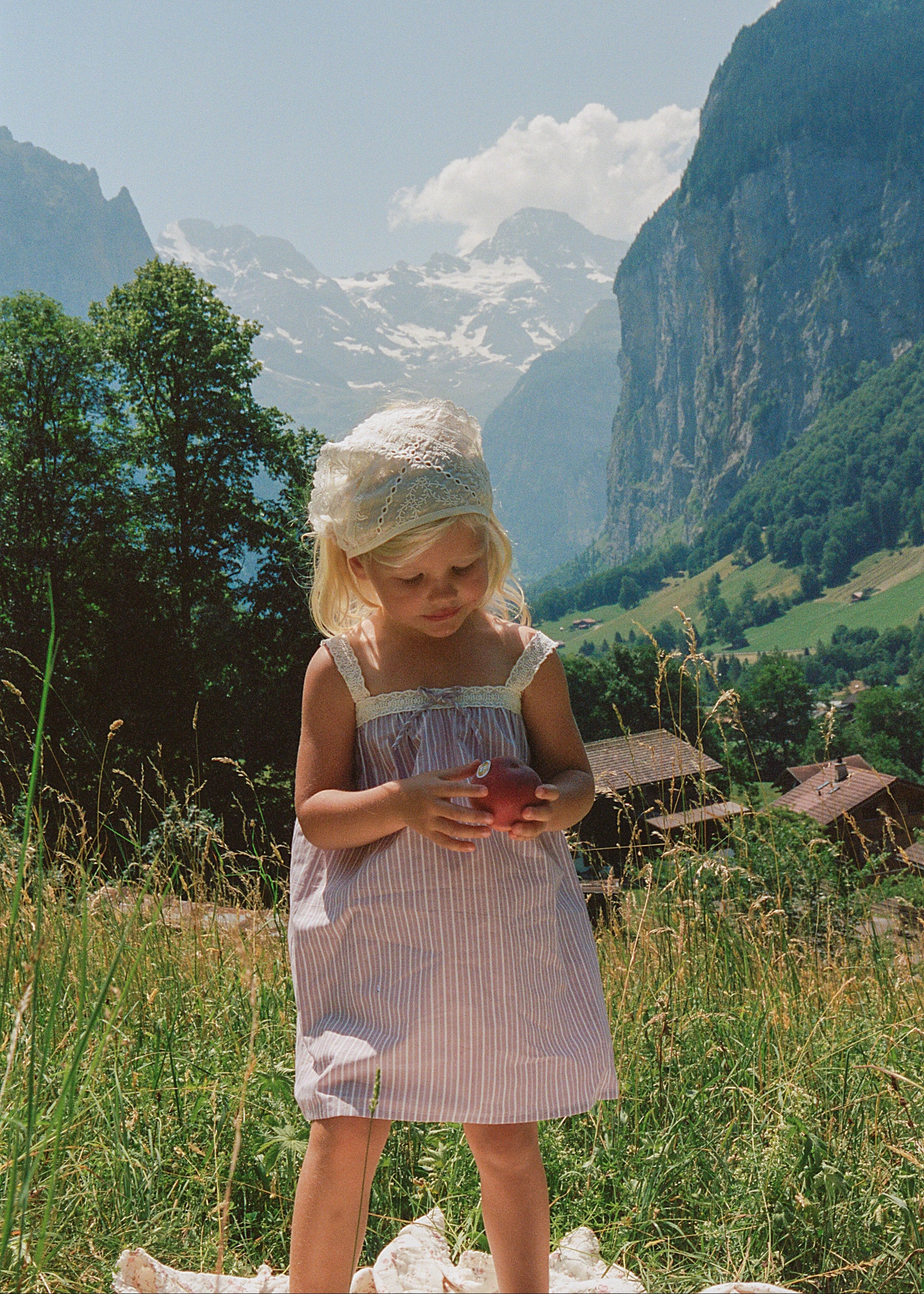 A young girl in a light pink and white striped dress and white bonnet stands in a grassy mountain valley holding a red apple, with steep cliffs and snow-capped peaks in the background.