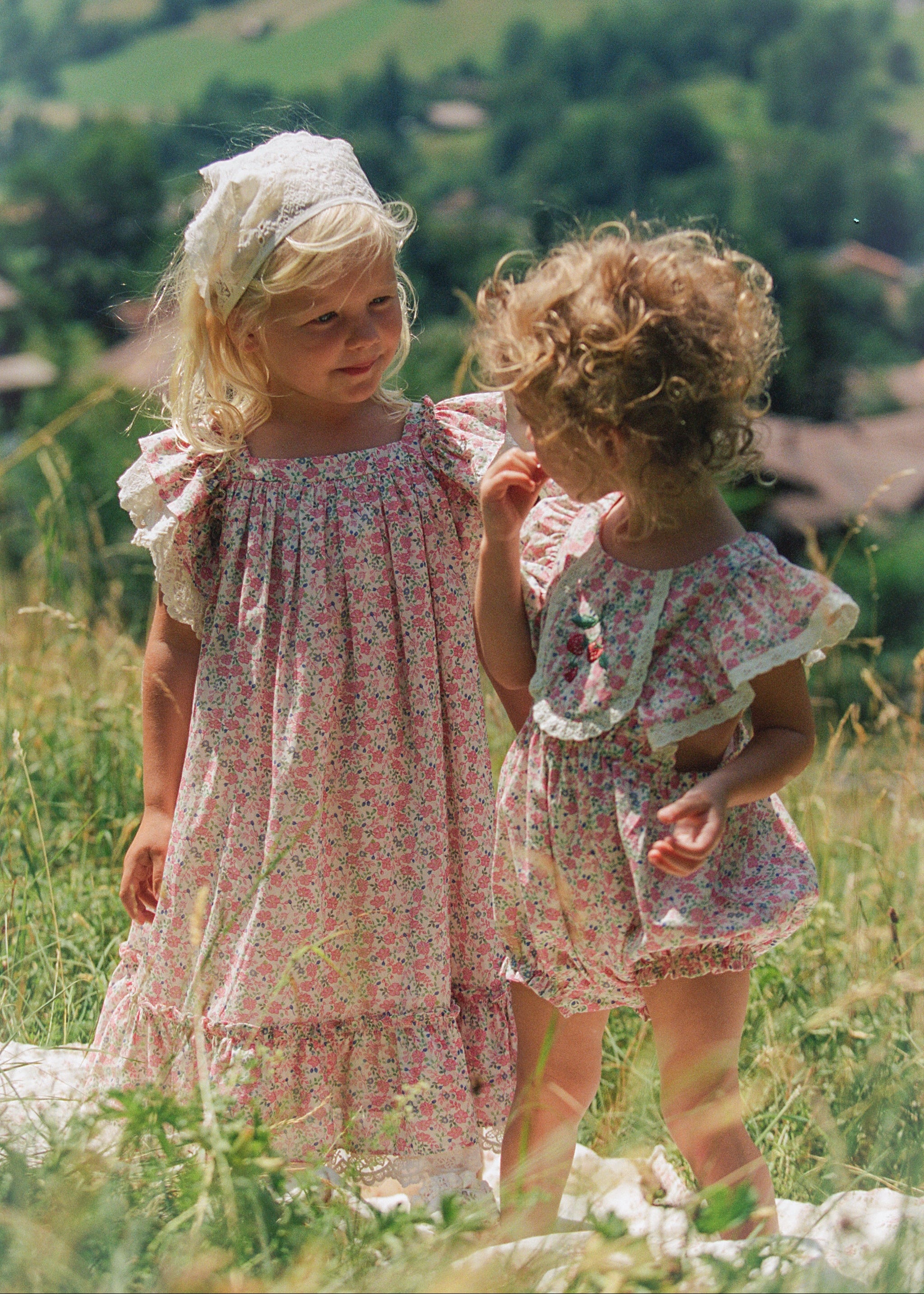 Two young girls standing outdoors in a grassy field, wearing matching floral dresses. The background shows a blurred mountain landscape.