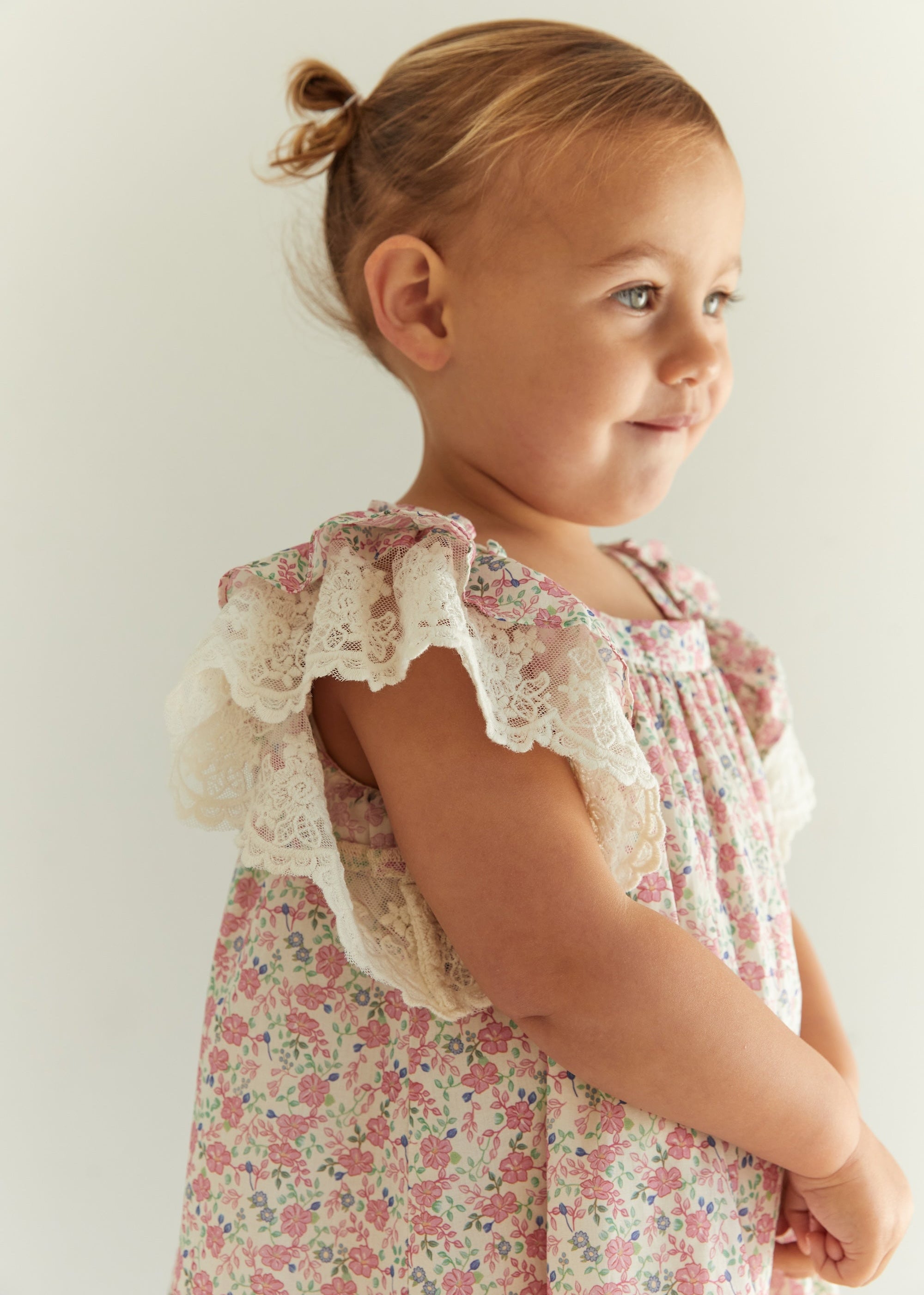 A young toddler girl with blonde hair in a small bun, wearing a pink and white floral print dress with cream lace ruffle shoulder details, photographed against a plain light background.
