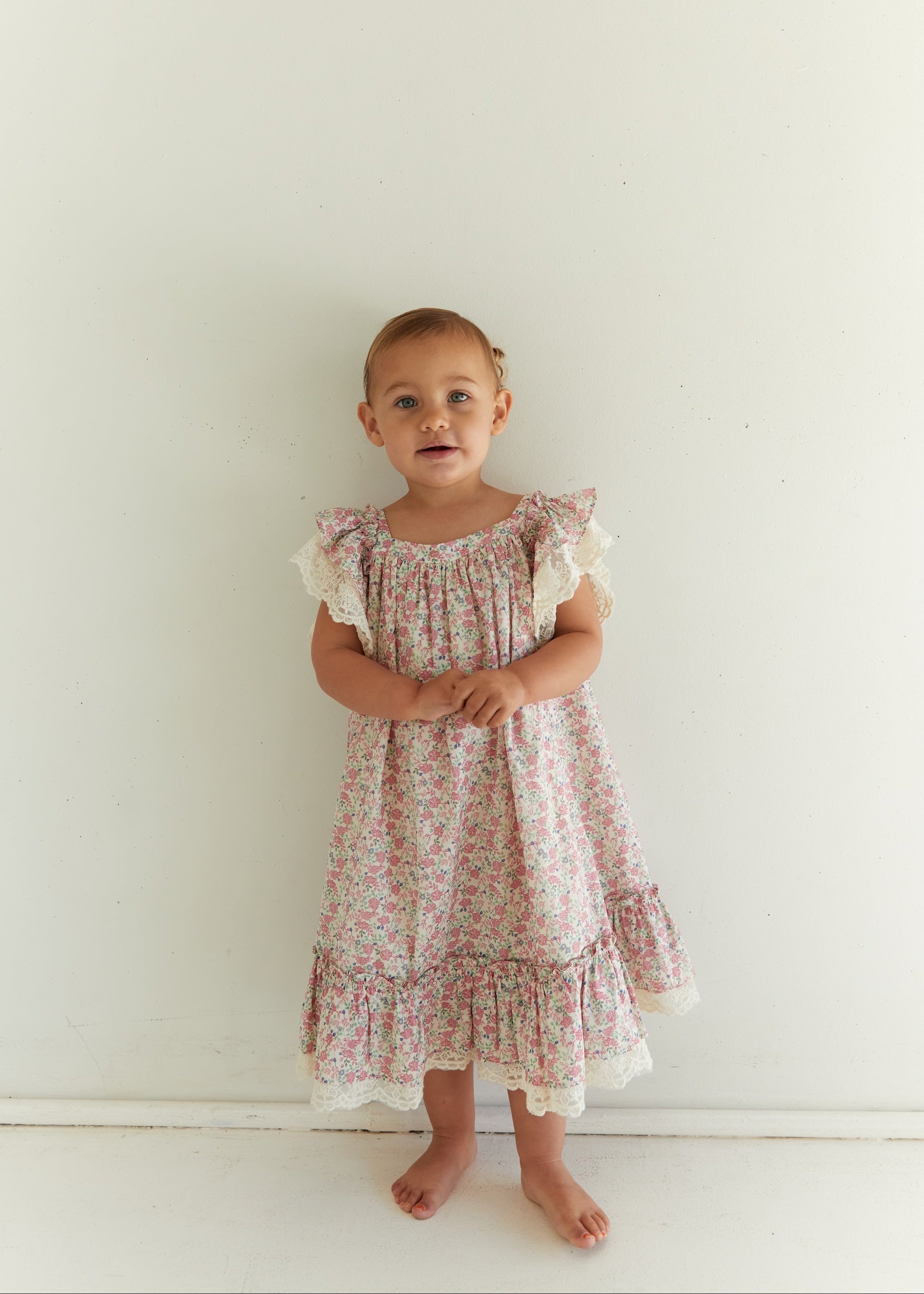 A toddler girl stands barefoot against a plain white wall wearing a light pink and white floral print dress with ruffled sleeves and a ruffled hem.