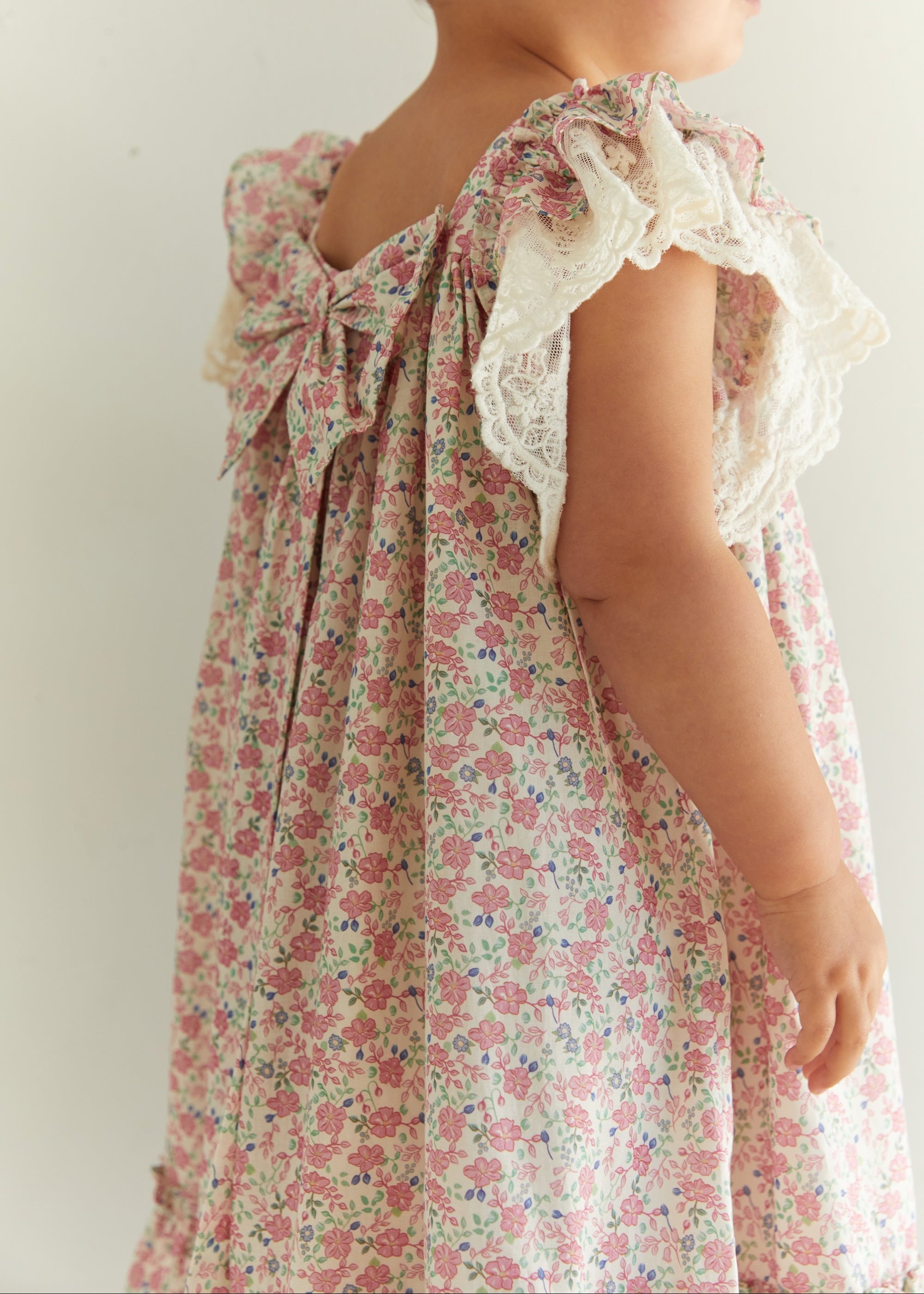 A child wearing a floral print nightgown with ruffled lace shoulder straps and a large bow on the back, shown from the side against a plain light background.