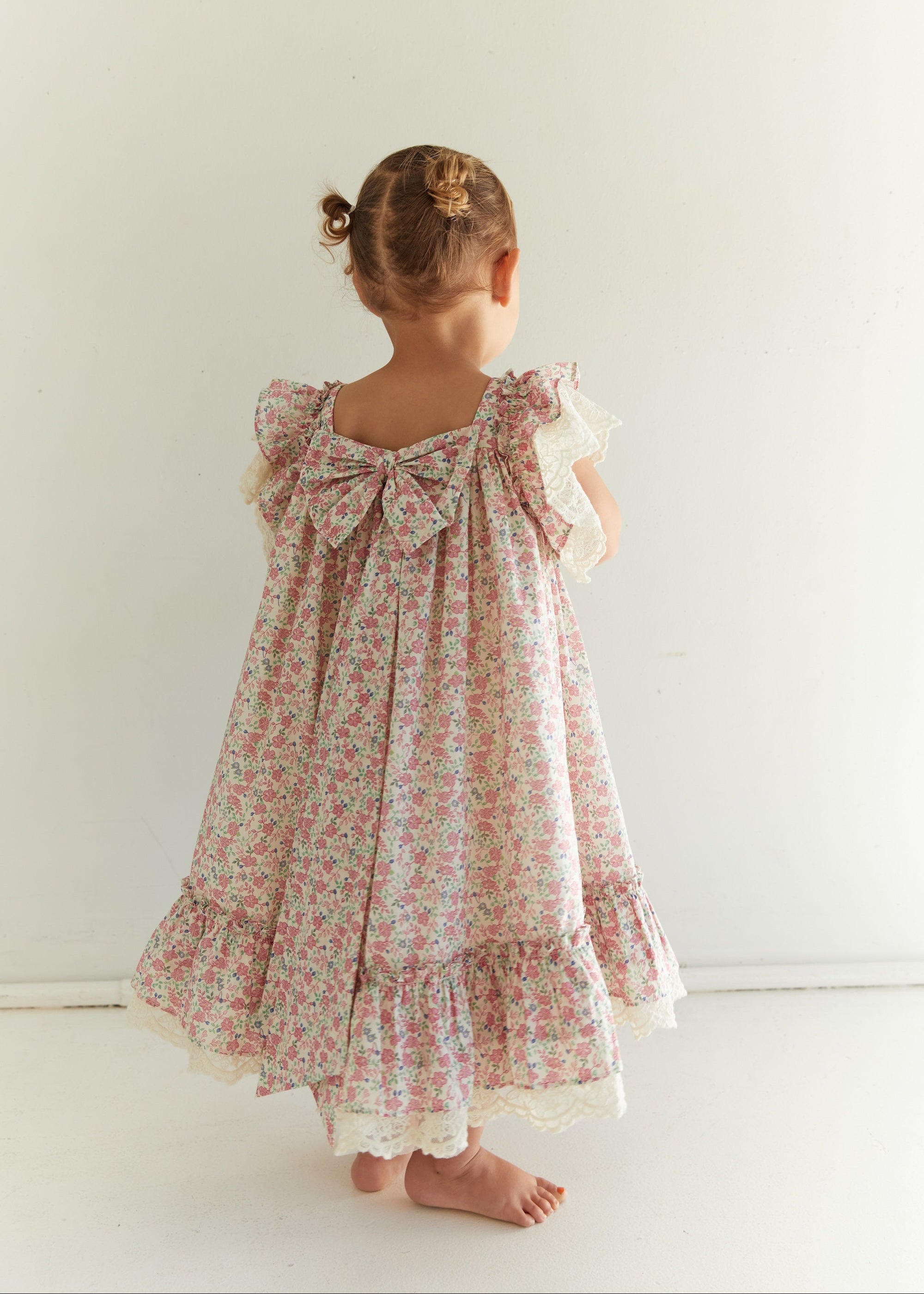 A young child viewed from behind wearing a floral print dress with a large bow at the back, ruffled sleeves, and lace trim, standing against a plain white wall.
