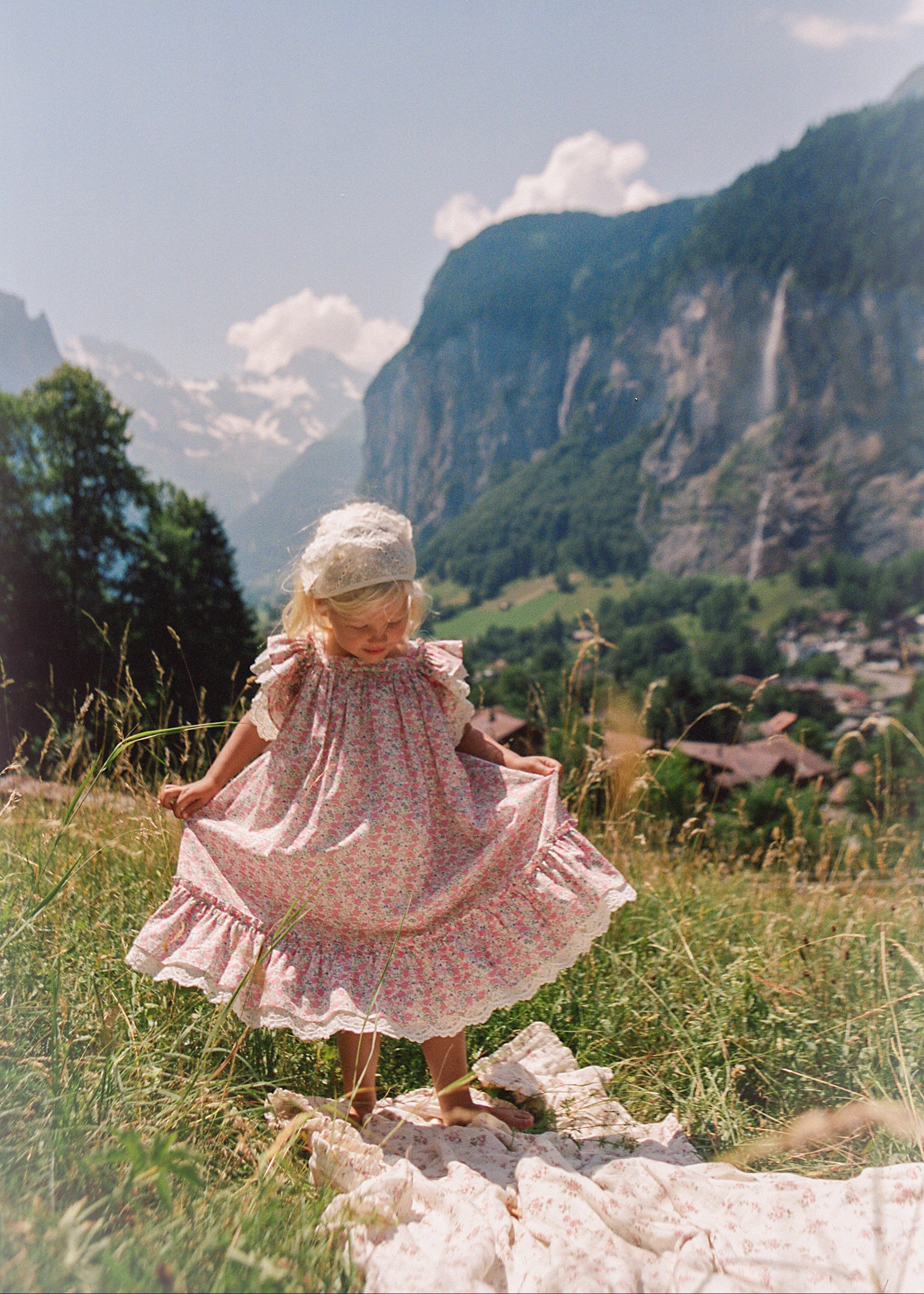 A young child wearing a pink floral dress and a white bonnet stands barefoot in a grassy field, holding out the sides of the dress to show its full patterned fabric, with a scenic mountain landscape in the background.