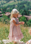 A young girl in a pink floral dress and white bonnet stands in a grassy field with a mountain village in the background.
