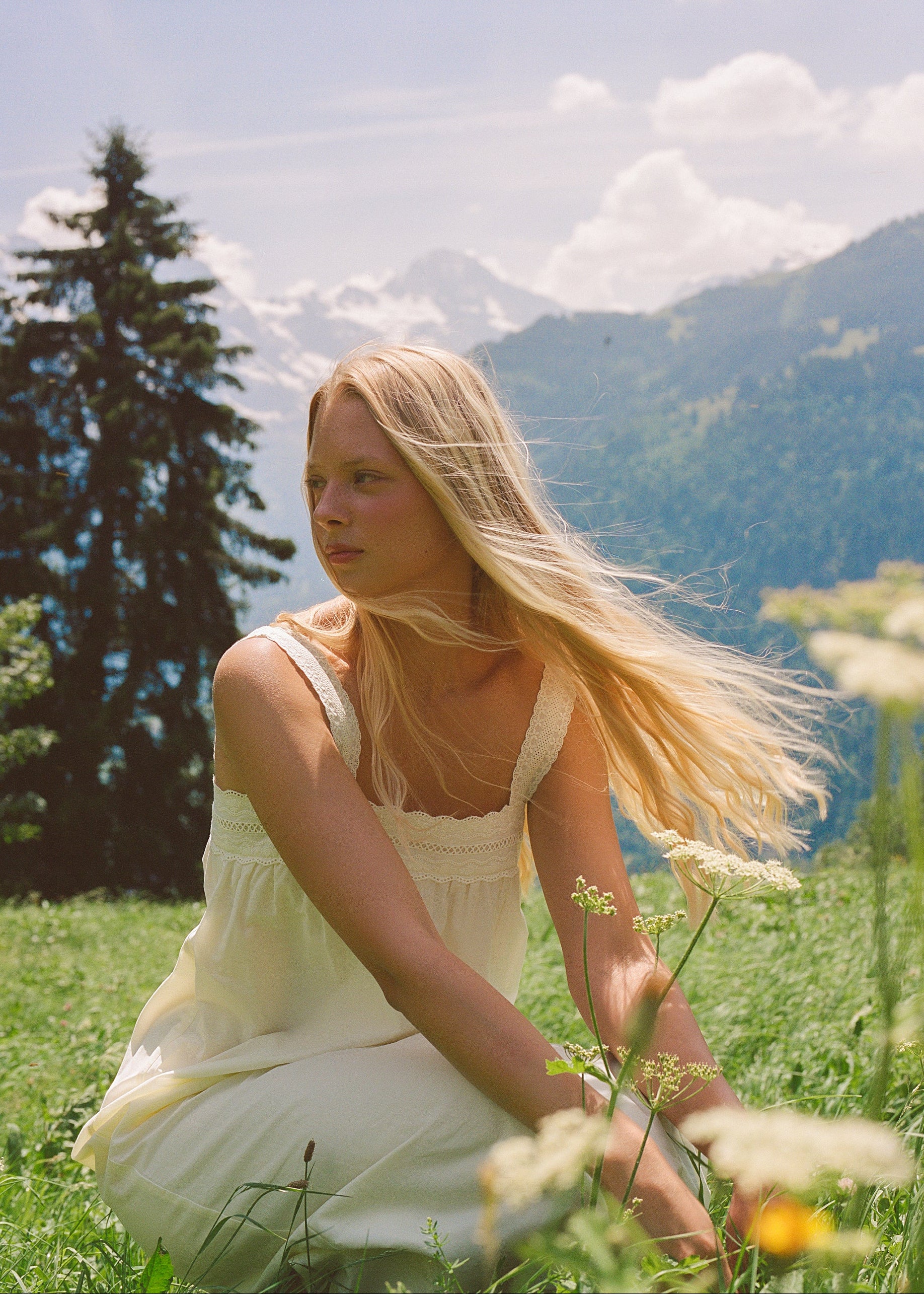 A young blonde girl wearing a cream-colored sleeveless dress sits in a grassy field with wildflowers, with mountains and pine trees in the background under a cloudy sky.