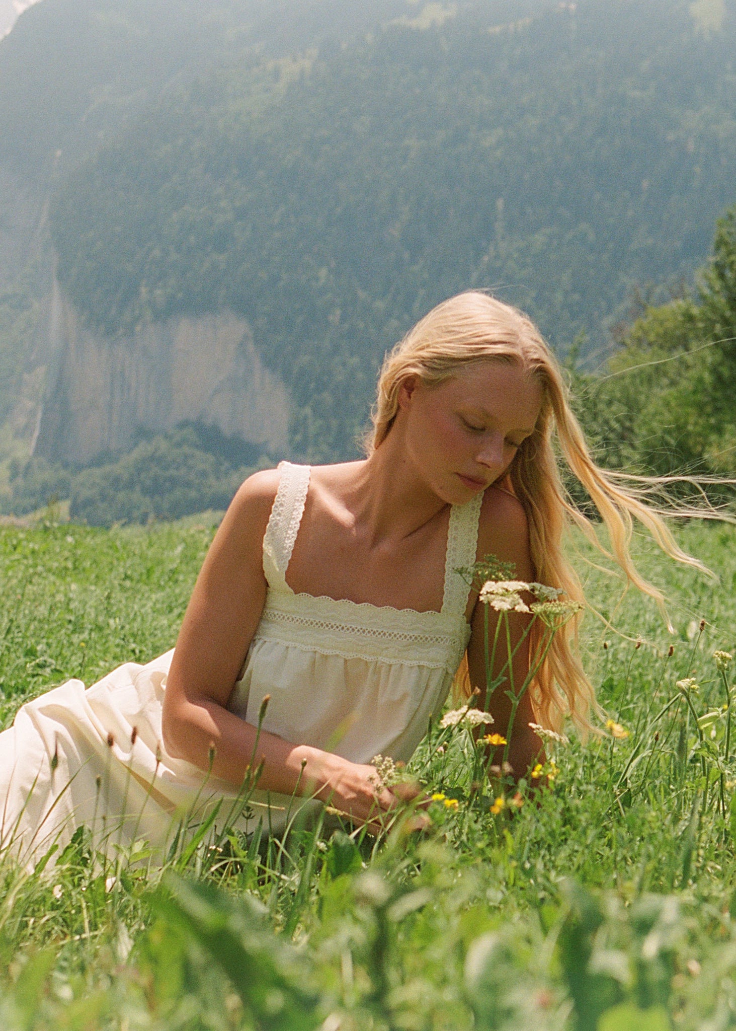 A woman in a cream-colored sleeveless dress lies in a grassy mountain meadow, looking down at wildflowers. A small wooden cabin and dense green forest are visible in the background, with a dramatic cliff face further behind her.