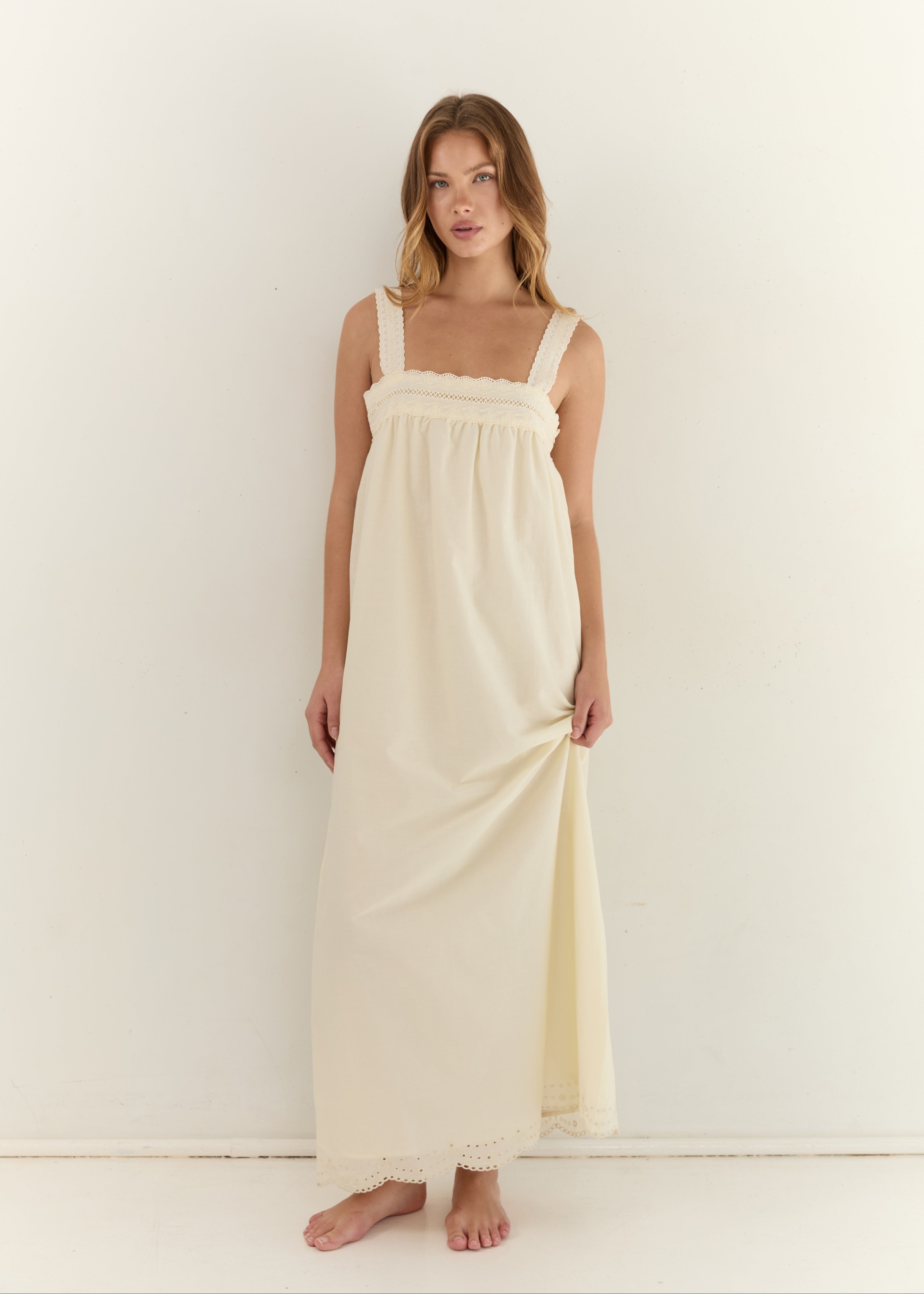 A woman modeling a long cream-colored cotton nightgown with a gathered bodice and scalloped edges, standing against a plain white wall.