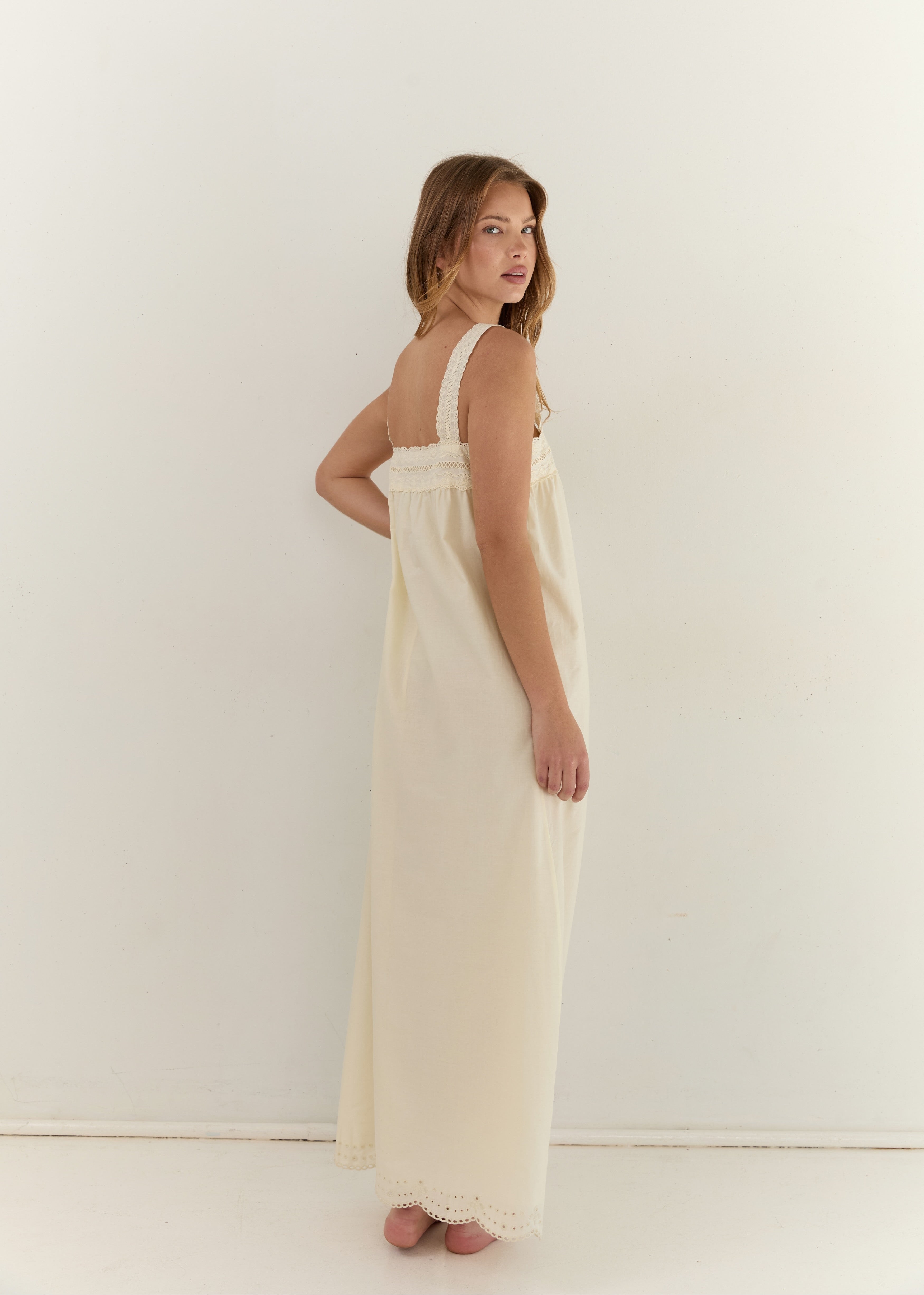 A woman wearing a long cream-colored slip dress with lace trim, standing against a plain white wall.