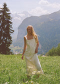 A woman with long blonde hair stands in a grassy mountain meadow wearing a white sleeveless dress, with snow-capped mountains and trees in the background.