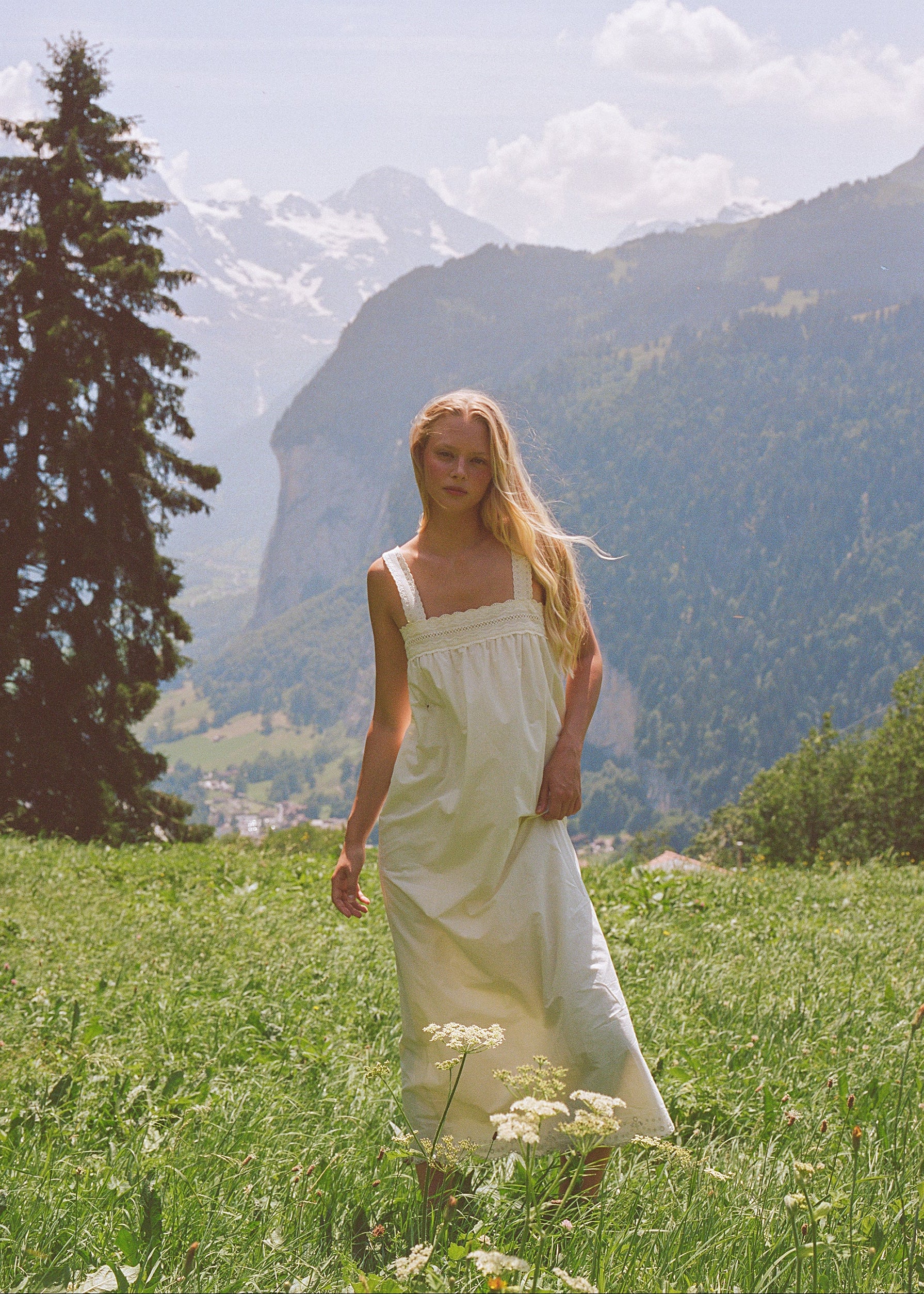 A woman with long blonde hair stands in a grassy mountain meadow wearing a white sleeveless dress, with snow-capped mountains and trees in the background.