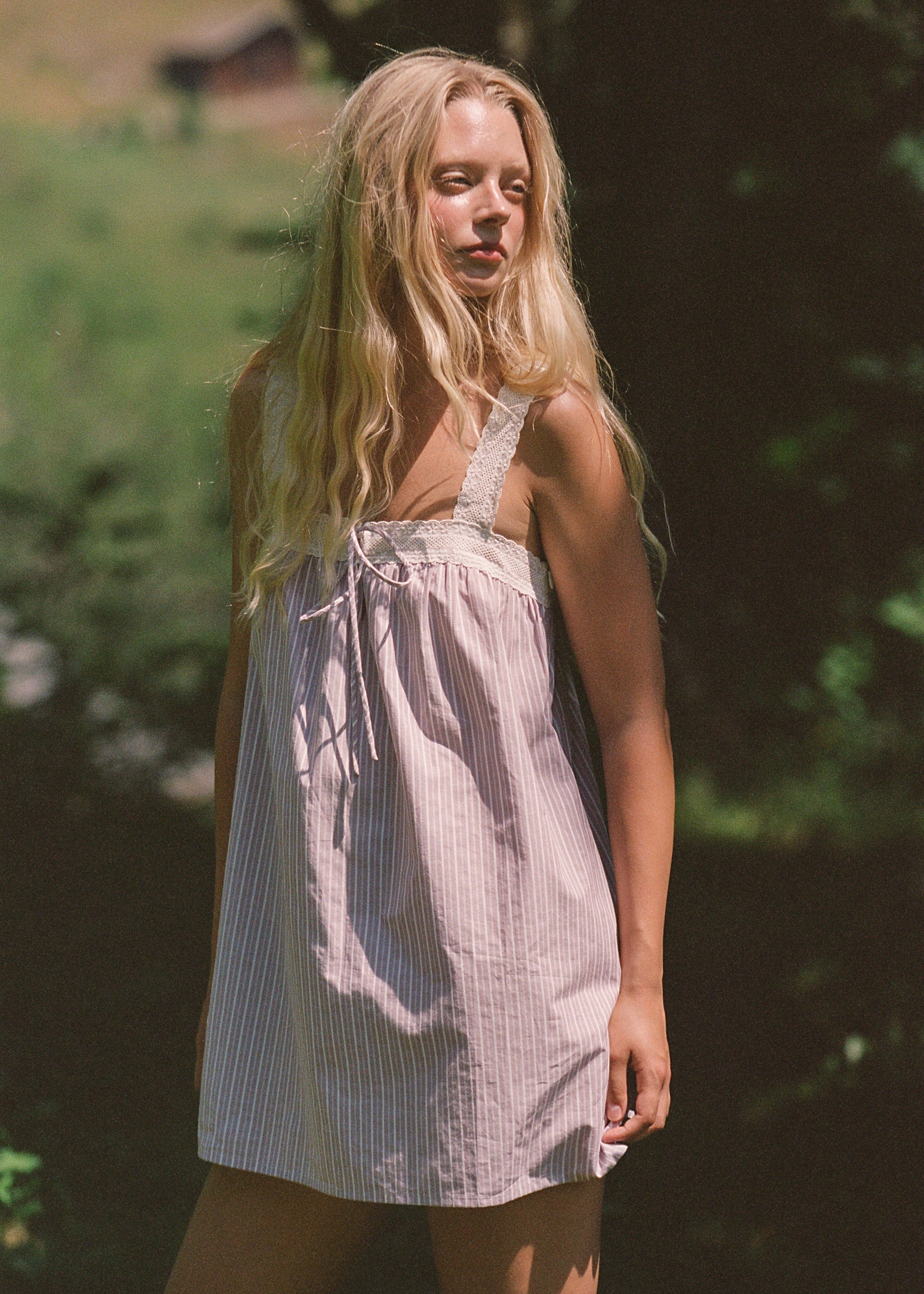 A young woman with long blonde hair stands outdoors in a wooded area wearing a light pink and white vertically striped sleeveless dress with lace shoulder straps.