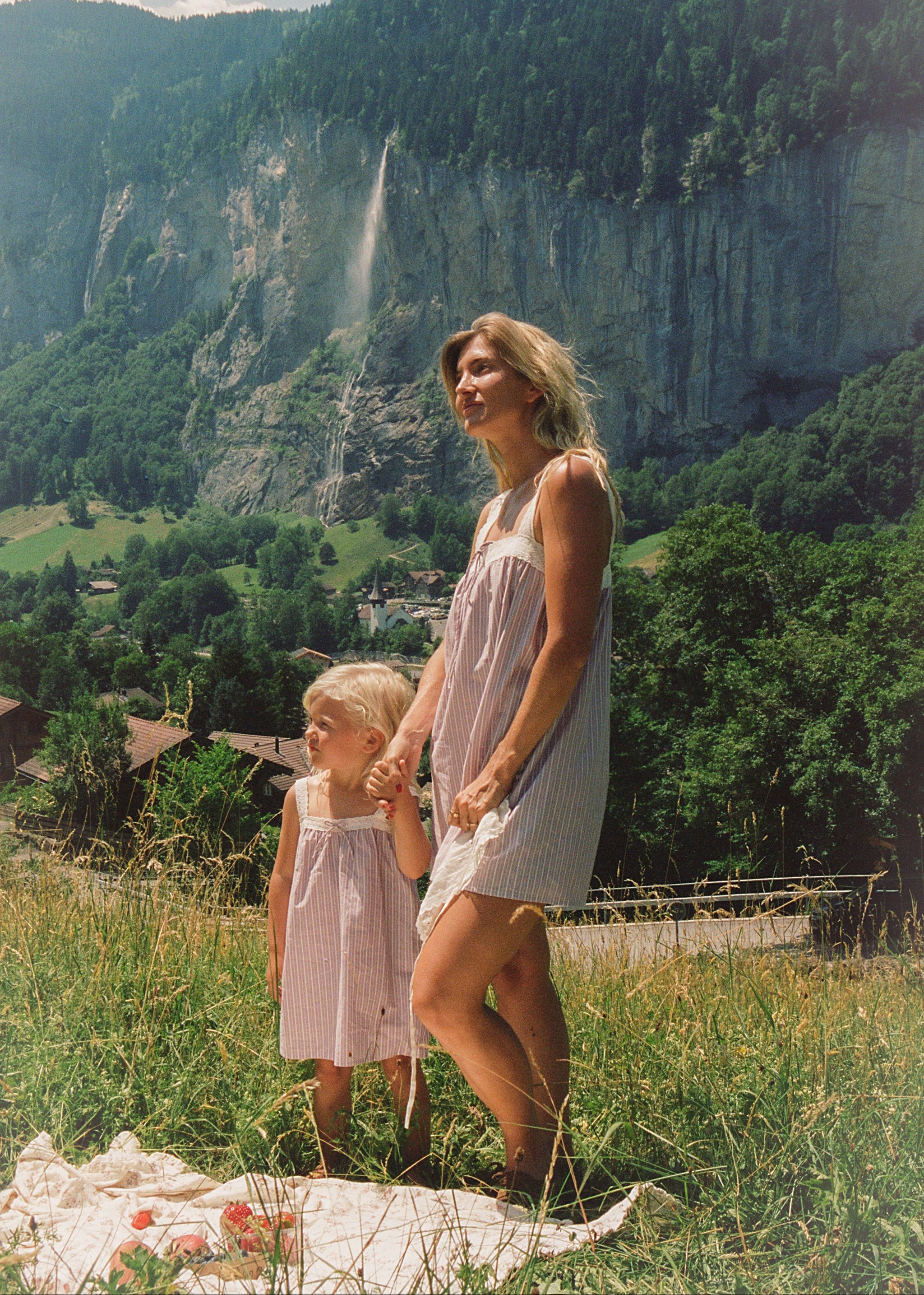 A woman and a young child wearing matching light pink and white striped sleeveless dresses stand on a grassy hillside holding hands, with a tall forested cliff and waterfall in the background.