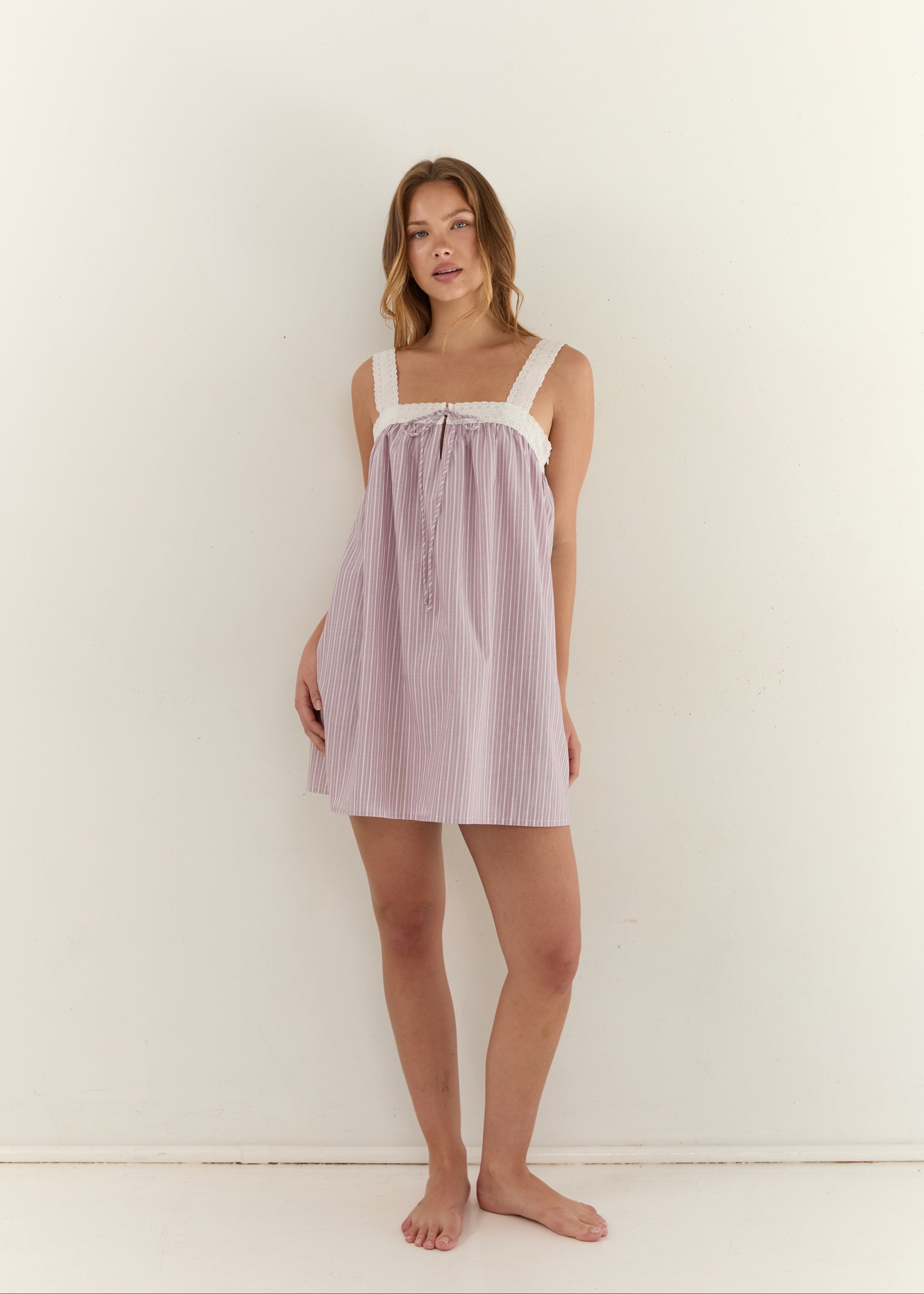 A woman modeling a light pink and white vertically striped nightgown with white lace trim on the straps, standing against a plain white wall.