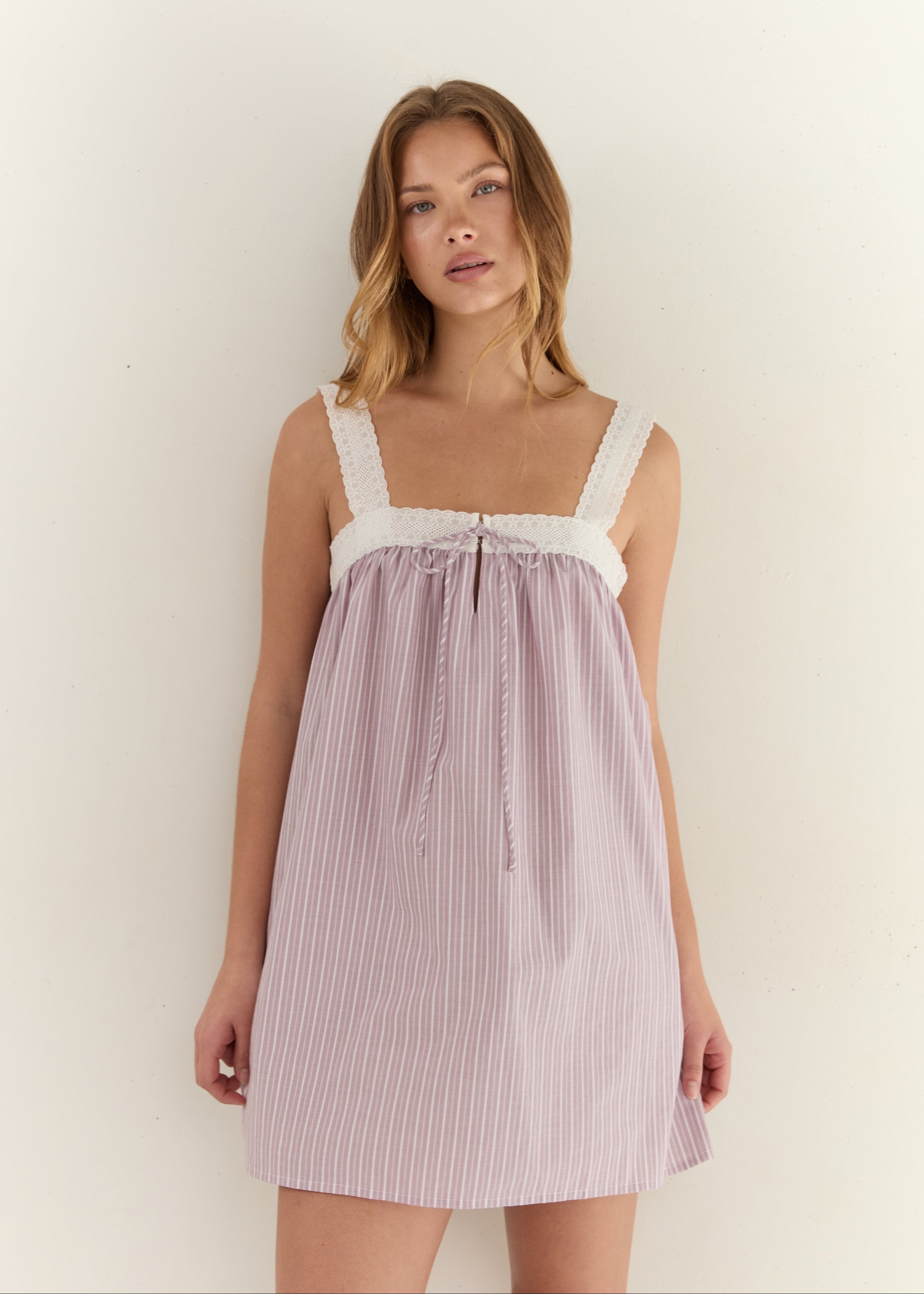 A woman modeling a light pink and white vertically striped nightgown with white lace trim on the straps and a gathered tie at the bust, set against a plain off-white background.