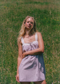 A woman stands in a grassy field wearing a light lavender and white vertically striped sundress with white lace trim on the straps and bust.