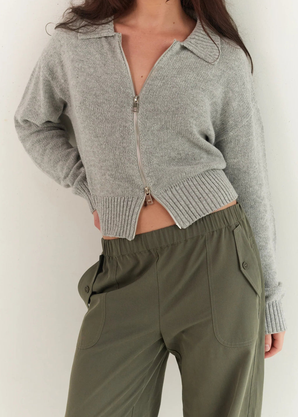Person wearing a gray zip-up cardigan and green pants against a white background