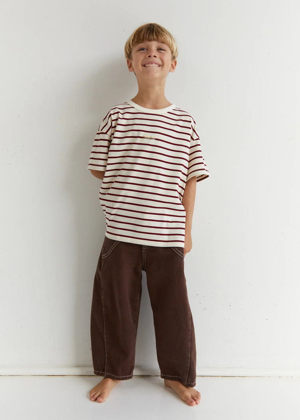 Child wearing a striped shirt and brown pants standing against a white wall.