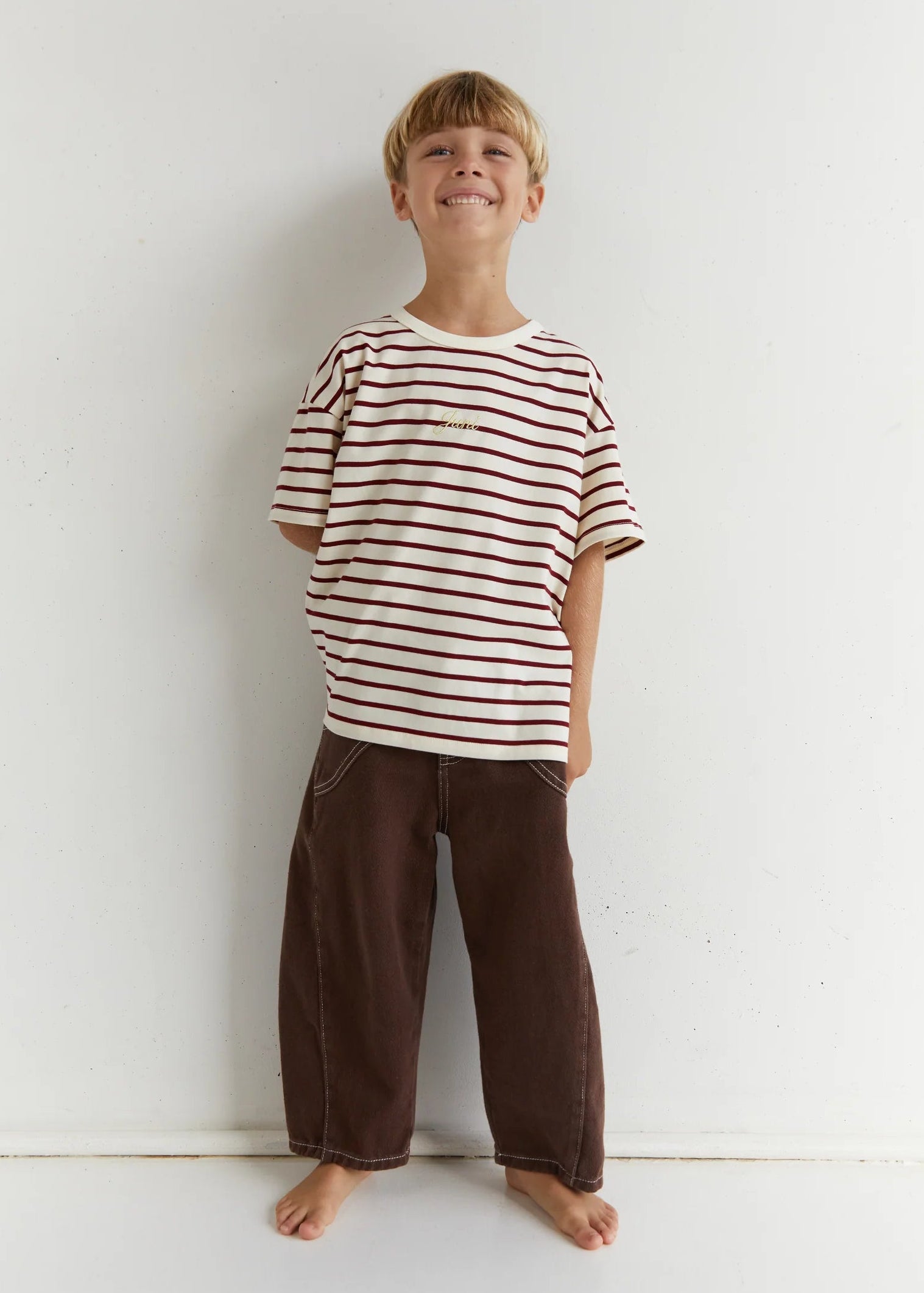 Child wearing a striped shirt and brown pants standing against a white wall.