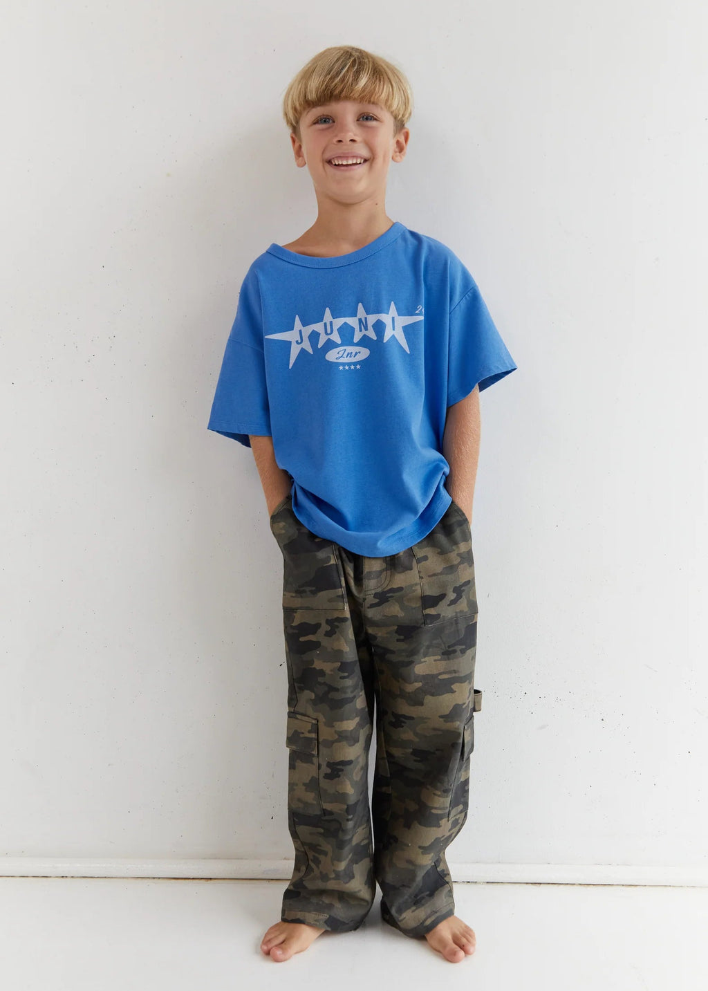 Child wearing a blue t-shirt with a logo and camouflage pants against a white background
