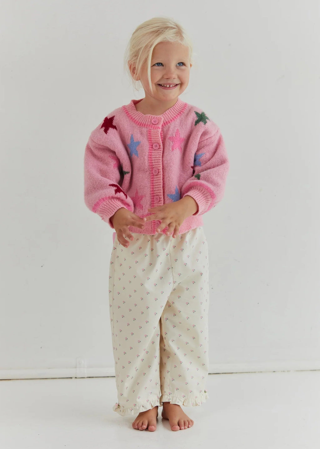 Young girl wearing a pink cardigan with colorful buttons and white pants with a pattern, standing against a plain background.