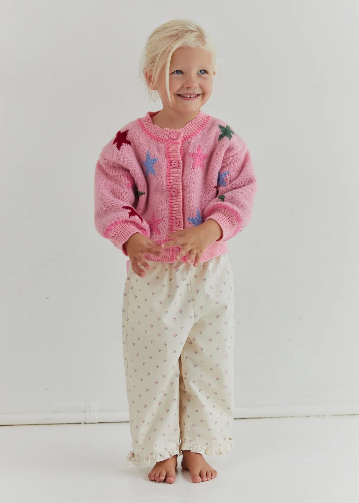 Young girl wearing a pink cardigan with colorful buttons and white pants with a pattern, standing against a plain background.