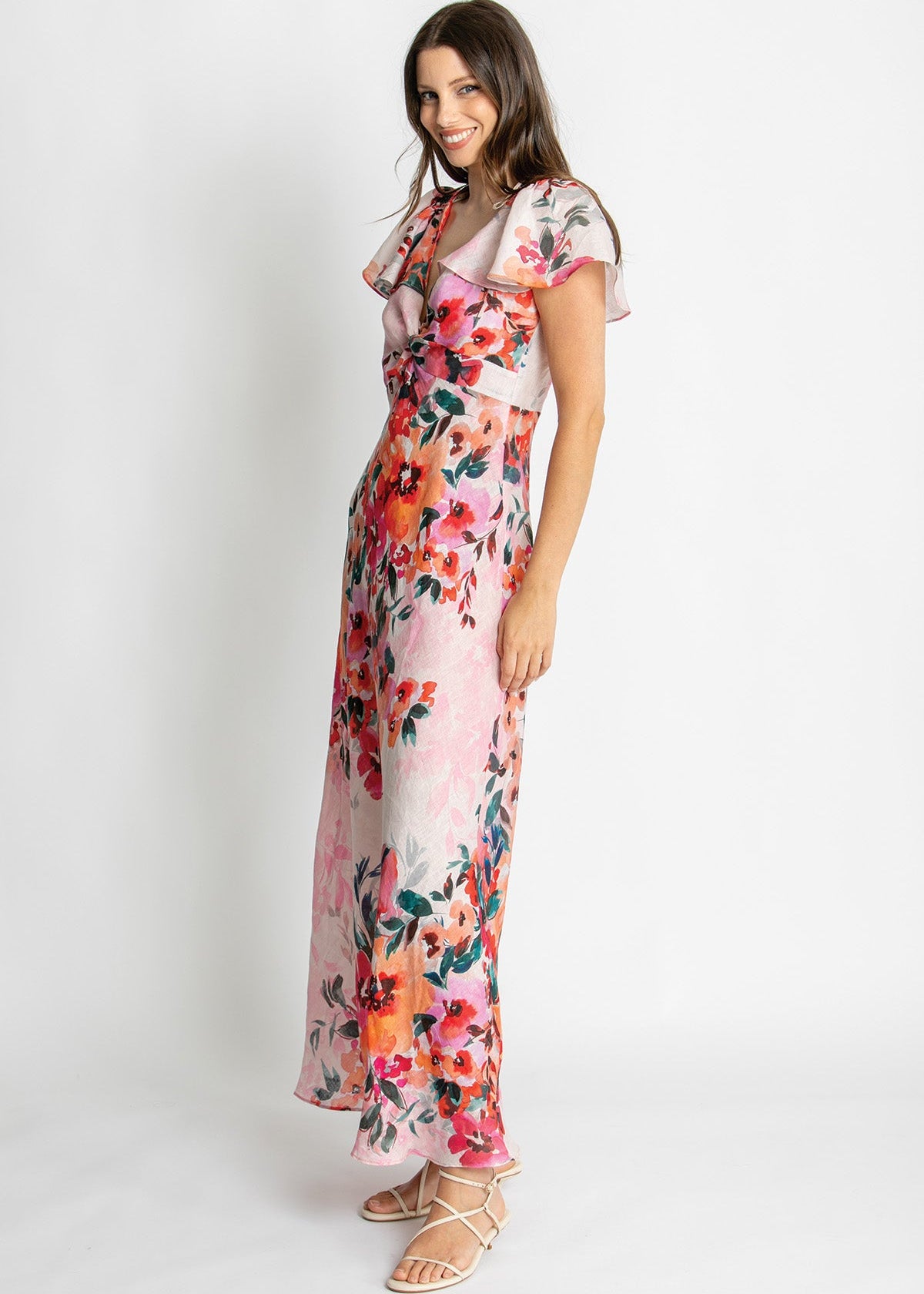 A woman modeling a long pink floral maxi dress with ruffled short sleeves and a gathered waist, paired with beige strappy sandals against a plain white studio background.